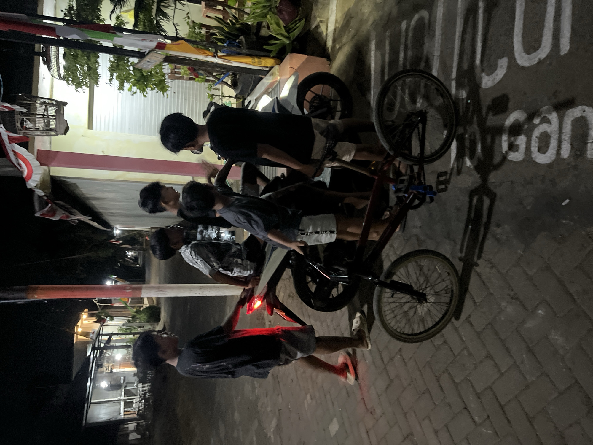 In Karangrandu, Indonesia, at night, four young males are gathered around a motorcycle and a bicycle. One male on the left, wearing a dark t-shirt and light shorts, stands near the rear of the motorcycle, with its brake light illuminated red. He is facing away from the viewer. To his right, two other males are partially visible, one in a patterned shirt and another in a dark shirt. The fourth male, on the far right, is also in a dark shirt and light shorts, positioned on the other side of the motorcycle. A bicycle is in the foreground, with its front wheel closest to the camera. The scene takes place on a brick-paved pathway next to a building with a light-colored facade and a dark window. Flags are visible on the building, and a birdcage hangs from an overhang. The ground is marked with white lettering, partially obscured by shadows.