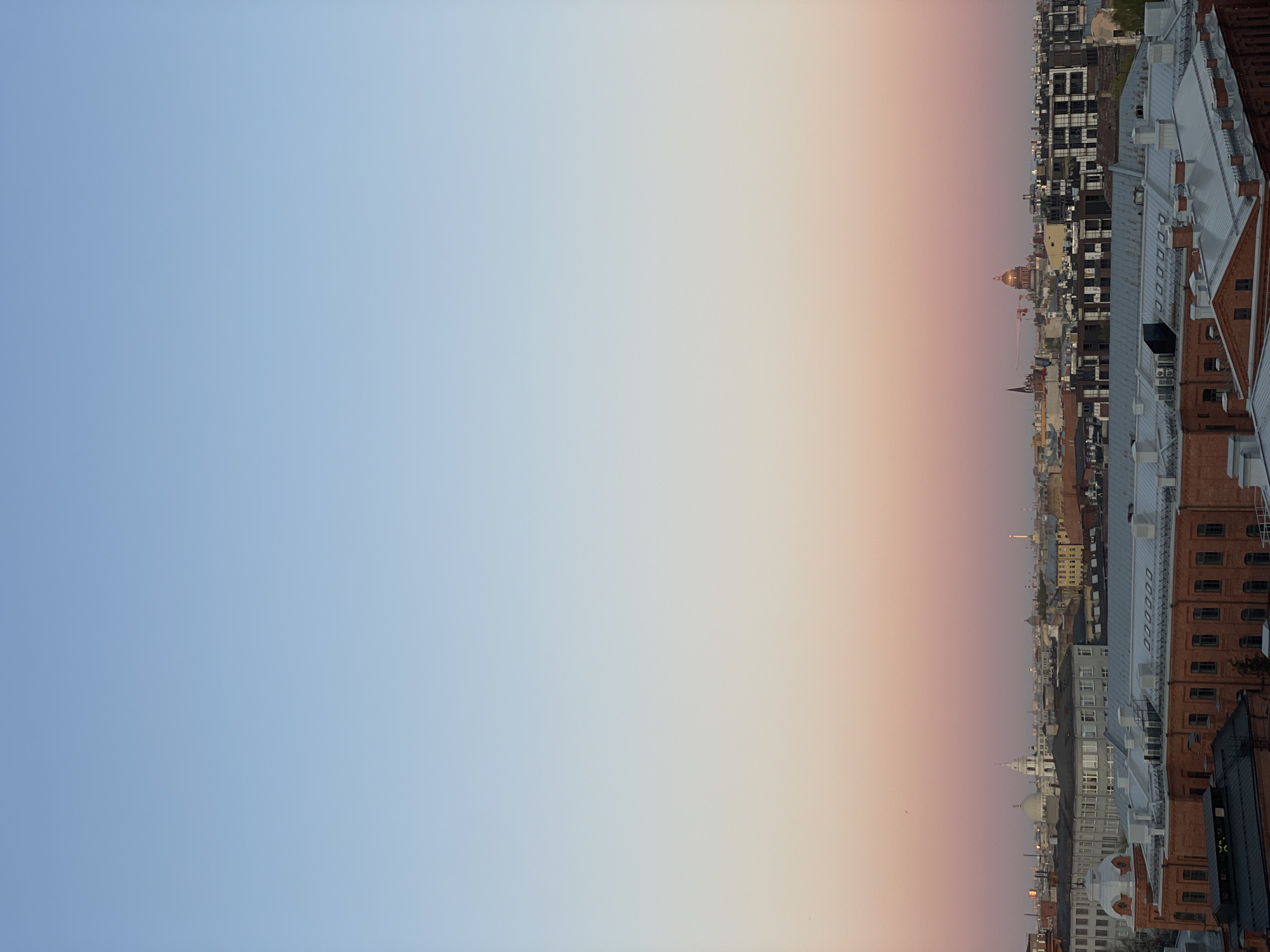 The image shows an upside-down view of Saint Petersburg, Russia, at dusk. The sky transitions from blue on the left to orange and pink towards the right. The cityscape on the right features rooftops, buildings of varying heights and architectural styles. Notably, there's a golden dome visible among the buildings. The light suggests a sunset or sunrise.