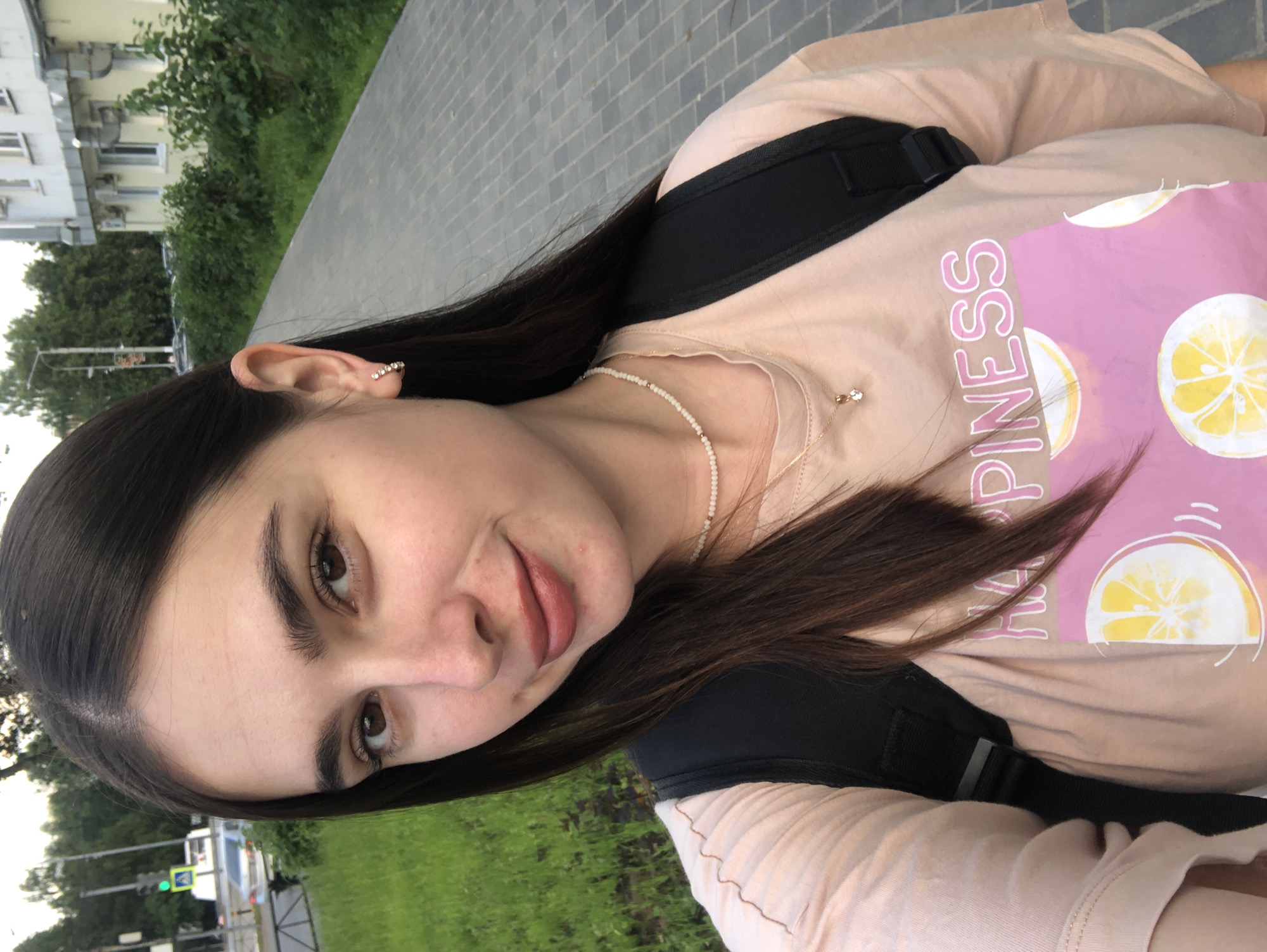 The media file is a portrait of a young woman. She has long, dark brown hair, brown eyes, and fair skin. She is wearing a light pink t-shirt with a graphic of lemons and the word "HAPPINESS" printed in pink. She also wears a delicate white beaded necklace and an earring with small gems. A black backpack is visible over her shoulders. The woman is looking slightly to her left, with a neutral expression.
The setting appears to be outdoors, likely on a sidewalk or path next to a grassy area. In the background, there is a building with several windows and air conditioning units, and trees with green foliage. A street with a traffic light and a car is also visible in the distance. The lighting suggests it is daytime, possibly overcast or late afternoon, given the soft light. The overall mood is casual and relaxed.
The visible text on her shirt reads "HAPPINESS". The location context is Chaikovsky, Russia.