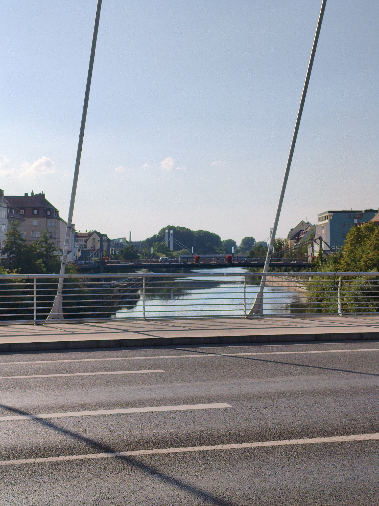 A wide-angle, eye-level, horizontal outdoor shot depicts the Regnitz river in Bamberg, Germany, under a clear blue sky with scattered white clouds. The foreground features an asphalt road with white lane markings, and the midground shows a pedestrian bridge with a silver railing and two tall, slender white poles supporting the bridge's structure. Beyond the pedestrian bridge, the Regnitz river flows, reflecting the sky and surrounding buildings. Several structures and trees line the riverbanks, including multi-story buildings with various architectural styles and lush green trees. A darker, wider bridge spans the river in the distance, with cars visible crossing it. The overall scene is peaceful and urban, showcasing a typical day in Bamberg.