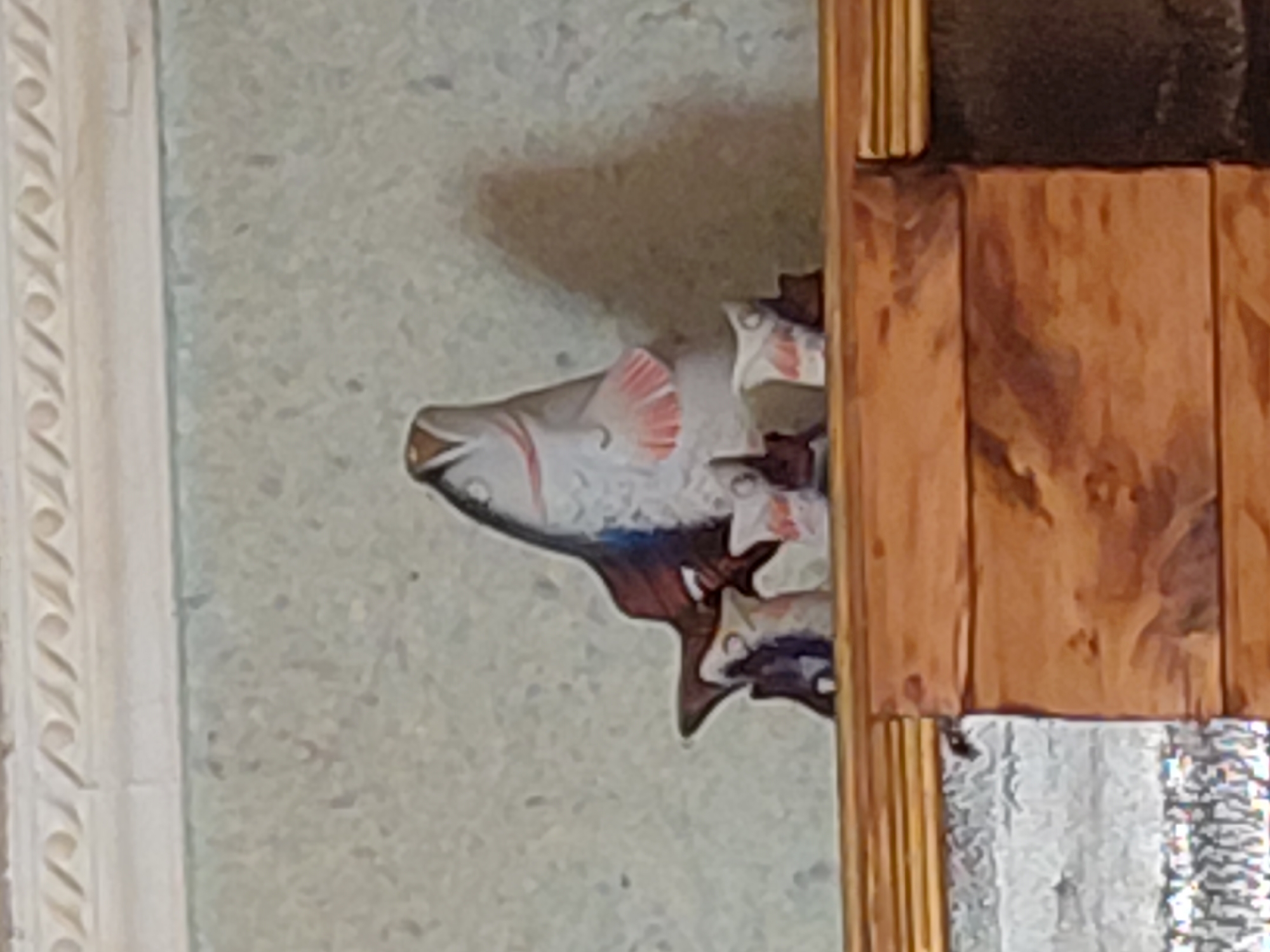 A kitschy, ceramic fish figurine with a gaping mouth and pink fins stands on a wooden shelf. The fish is positioned in the center of the shelf, with three small, cylindrical objects placed in front of it. These objects appear to be shot glasses or small cups, each with a white base and a red design. The background is a textured, light-colored wall, and a decorative molding runs along the top edge of the frame. The shelf itself is made of dark, polished wood. The scene is indoors, with the lighting suggesting it is daytime. There are no people or animals visible in the image. The location is described as Marynivka, Ukraine, though this information cannot be visually verified from the image alone.