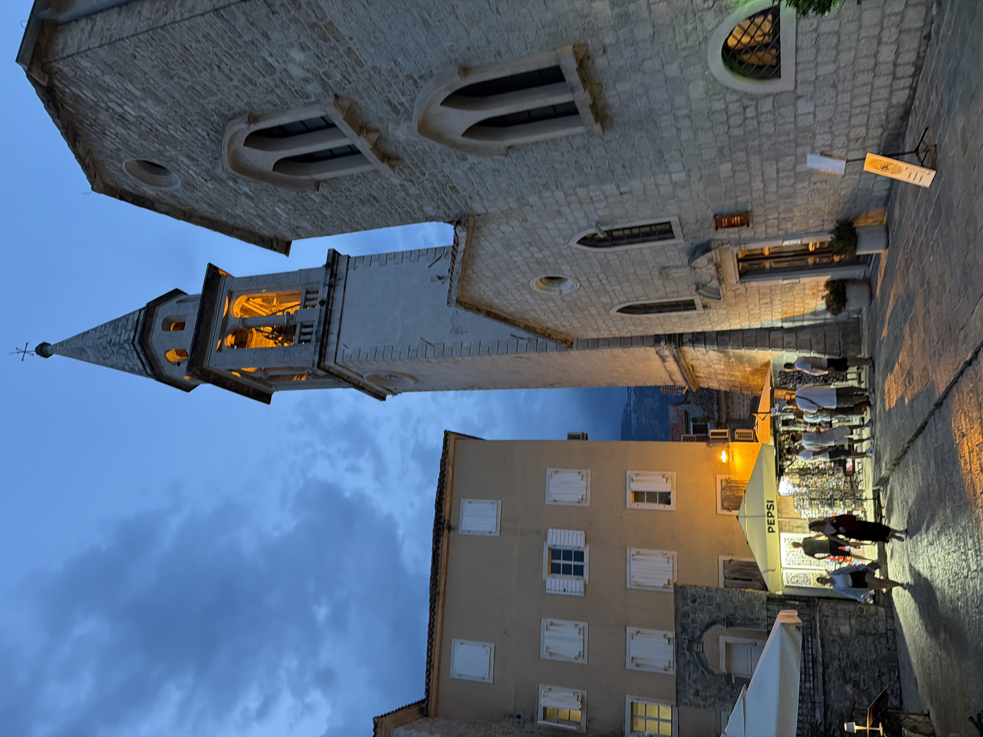 The image captures a street scene in Budva, Montenegro, at dusk. A prominent bell tower, illuminated from within, dominates the center of the frame, its peak reaching towards a cloudy blue sky. To the right, a stone building with arched windows suggests historical architecture, possibly a church or a part of the old town. To the left, a lighter-colored building with multiple shuttered windows stands, with a street-level shop front featuring a Pepsi-branded umbrella and display racks of merchandise. Several individuals are visible in the foreground and mid-ground, some walking along the cobblestone street, others browsing the shop. The lighting suggests artificial illumination from the shops and the bell tower, casting shadows on the ground. The overall atmosphere is that of a lively evening in a historic European town.