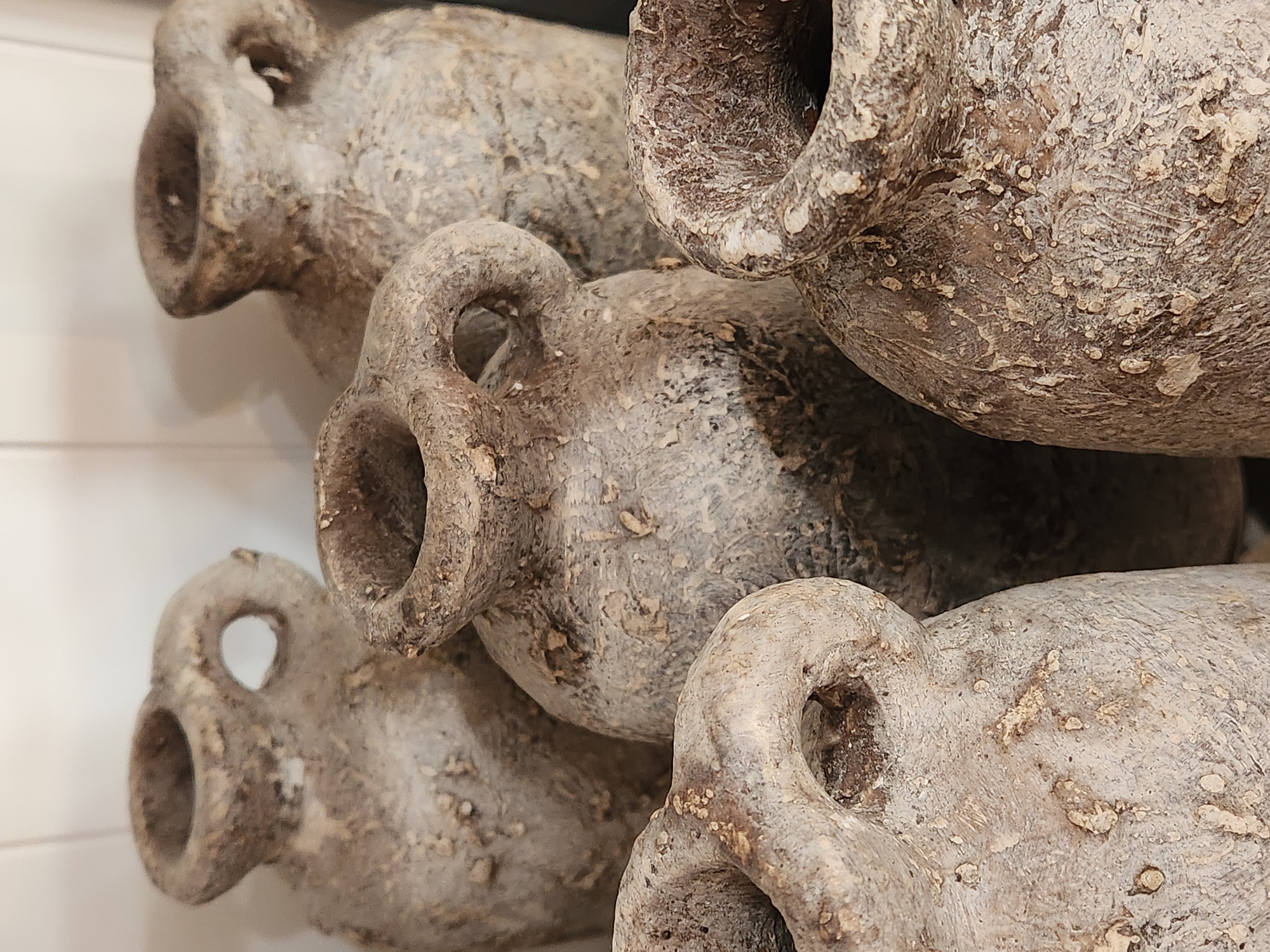 The image displays a close-up, angled view of a stack of weathered, ancient-looking amphorae. These are ceramic vessels with two handles and a narrow neck, often used for storage and transport in ancient times. The amphorae are arranged in a layered fashion, with their rims and handles partially obscuring each other.
The texture of the amphorae is rough and uneven, showing signs of age and wear. They have a predominantly grayish-brown hue with lighter, patchy areas that suggest salt encrustations, mineral deposits, or remnants of their original contents. The surfaces are not smooth but rather textured with small bumps and irregular patterns, contributing to their antique appearance.
The focus is on the objects themselves, with no people or clear environmental details like weather or time of day visible. The background is blurred, with a hint of horizontal lines on the left suggesting a wall or surface. The lighting appears to be soft and diffused, highlighting the texture of the amphorae without casting harsh shadows.
The arrangement and close-up perspective emphasize the form and material of these historical artifacts. They evoke a sense of history and antiquity, likely part of a museum display, a historical reenactment, or a curated collection. There is no visible text in the image. The context provided places this scene in Maitland, United States of America.