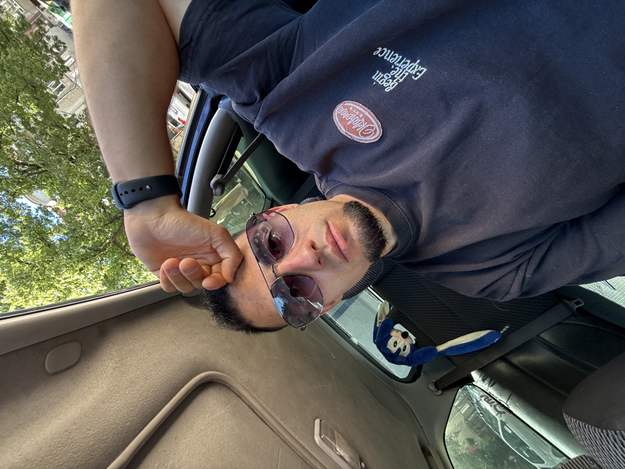 The image shows a close-up, high-angle shot of a man in a car, likely taken by himself. He is wearing sunglasses and a dark sweatshirt with a patch on the left chest that reads "Oklahoma" and text below it saying "Begin the Experience." His left arm is resting on his forehead. A blue stuffed toy is visible on the back seat. The car's interior, including the sunroof and part of the dashboard, is visible. Through the car window, trees and buildings can be seen, suggesting an urban or suburban setting. The overall lighting indicates it is daytime, possibly a sunny day, given the shadows and bright greenery.