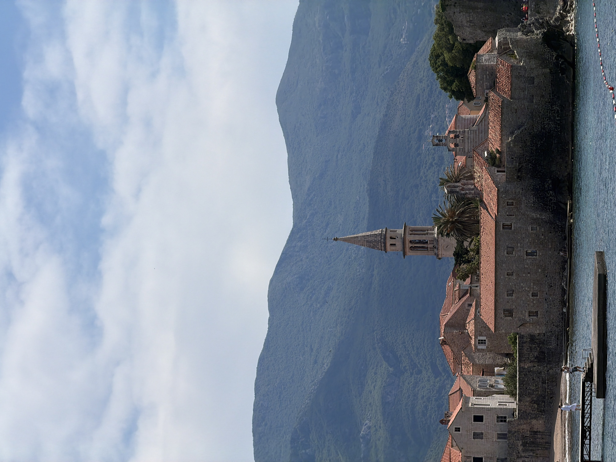 The image shows the Old Town of Budva, Montenegro, viewed from the water. Buildings with red tile roofs and stone walls line the coast, punctuated by the prominent bell tower of the Church of St. Ivan. A large, forested mountain forms the backdrop. In the lower right, a small pier with people standing on it extends into the water. The sky is blue with scattered clouds. A rope with red and white buoys is visible in the water, possibly marking a swimming area.
