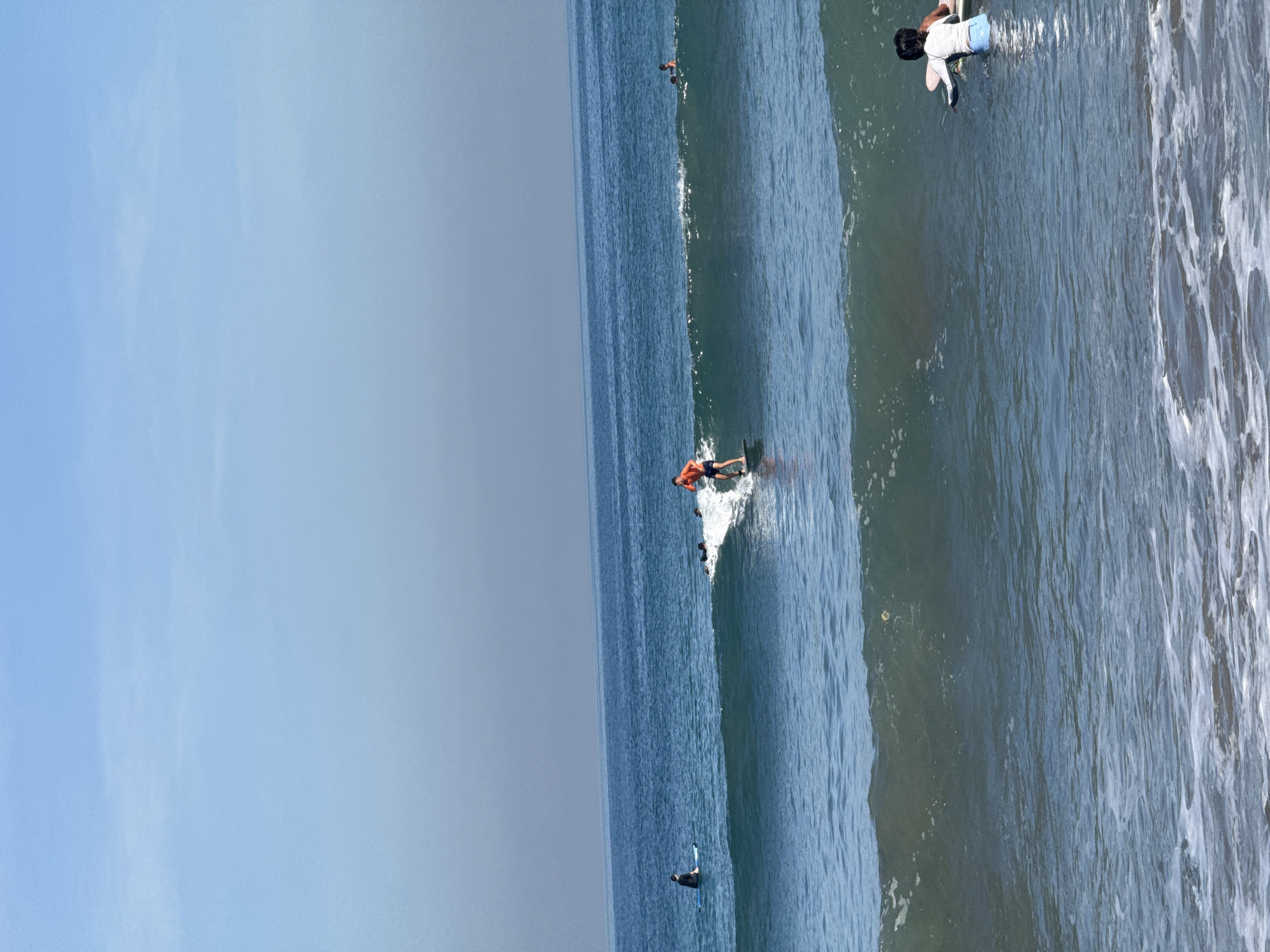 In Kuta, Indonesia, several surfers are in the ocean. The sky is a clear, pale blue, transitioning to a hazy grey near the horizon where it meets the sea. The ocean water is a deep blue, with visible waves breaking and rolling towards the shore.
In the foreground, a surfer in a white long-sleeved rash guard and blue shorts is sitting on a surfboard, facing away from the camera. Further out, a surfer in an orange top and blue shorts is actively riding a wave, standing on a surfboard. Several other figures, likely surfers, are scattered in the water, some partially submerged and others on their boards. One surfer in a black wetsuit is sitting on a light blue surfboard in the middle distance. The waves are moderate in size, with some white foam visible as they crest. The overall scene depicts a typical day of surfing on the coast of Kuta.