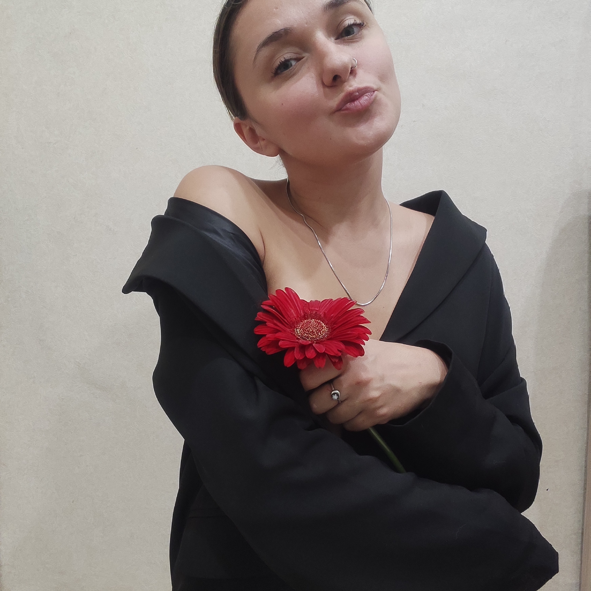 A young woman with her hair pulled back is posing indoors against a plain, light-colored wall. She is wearing a black blazer draped off one shoulder, revealing her bare shoulder and part of her chest. A silver necklace is visible around her neck, and a ring adorns her finger. She holds a single red gerbera daisy close to her chest with her right hand. Her facial expression is a slight pout. There is no visual information present that indicates the location in Yekaterinburg, Russia.