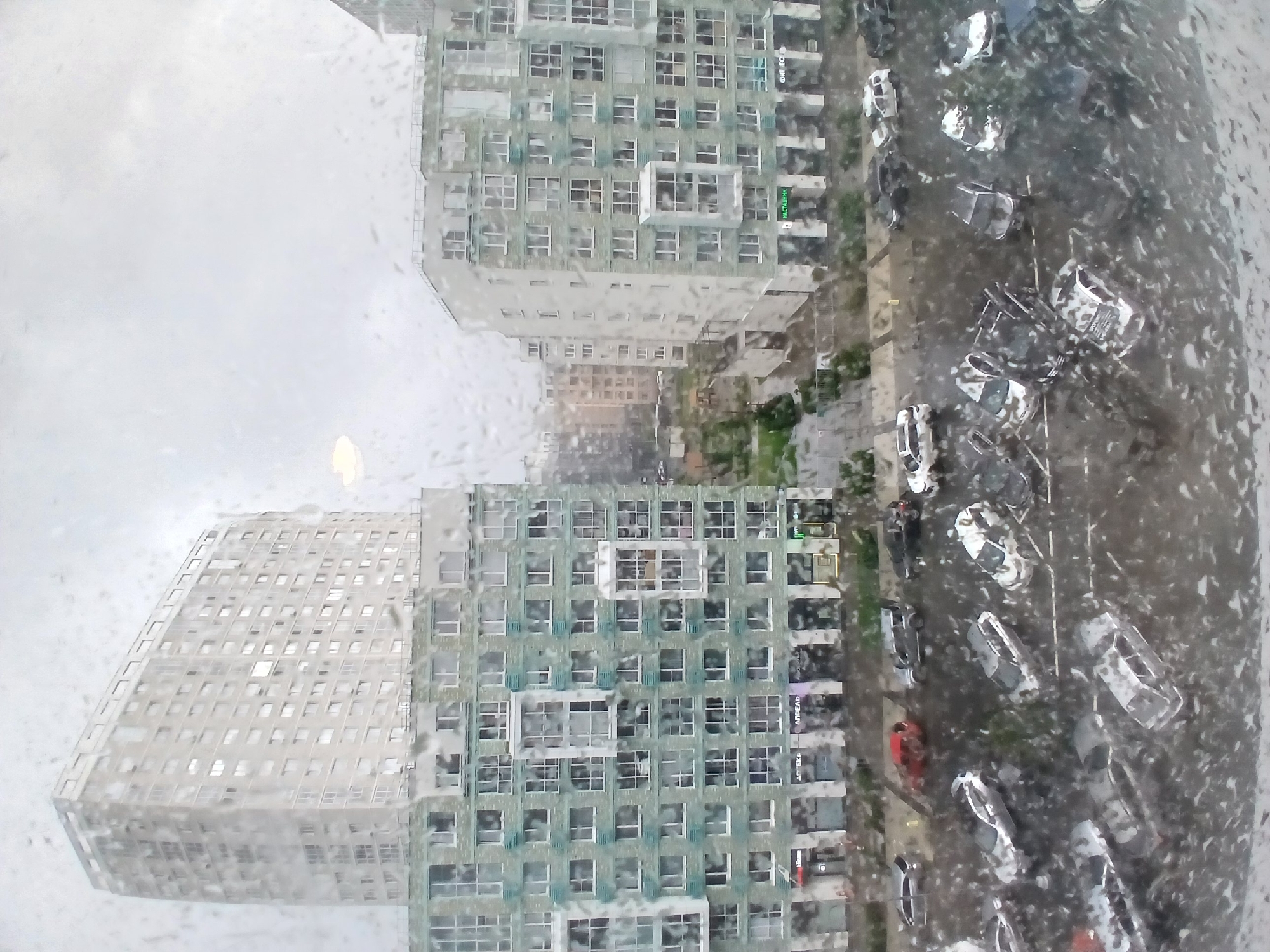 The image shows a view of a city street and buildings through a window with raindrops. On the left, a tall, light-colored residential building with many windows is visible. In the center and to the right, two modern, multi-story apartment buildings with light green facades and numerous windows are prominent. The lower floors of these buildings appear to house retail spaces with visible signage in Russian, including "АПТЕКА" (Apteka - Pharmacy) and "НАСТАВНИК" (Nastavnik - Mentor/Tutor). A street runs in front of these buildings, with several parked cars and a few cars in motion. The ground and cars appear wet, suggesting recent or ongoing rain. The overall scene depicts a typical urban environment, possibly during inclement weather. The style of the buildings and signage suggests this location is likely in Russia, and specifically Yekaterinburg, based on known architectural and commercial styles in the city.