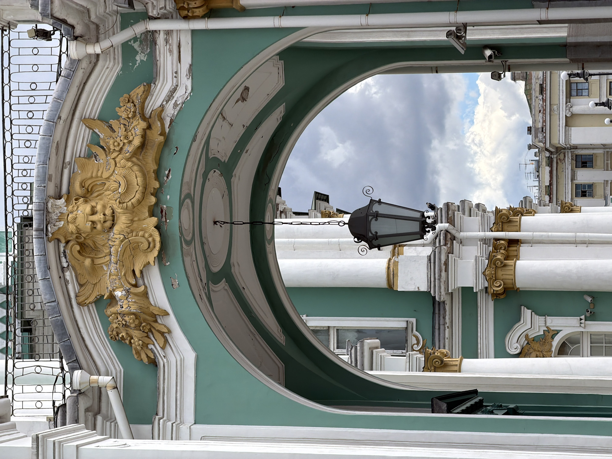 This media file is a photograph of the exterior of an ornate building, likely a historical palace or museum in Saint Petersburg, Russia. The image is oriented such that the sky is at the top and the ground is at the bottom.
The main subject is the architectural facade, characterized by its vibrant teal and white paint, extensive decorative stucco work, and classical columns. A large, circular opening, framed by elaborate molding, dominates the center of the image, offering a view of the sky and distant buildings. Through this opening, a dark, old-fashioned lantern hangs by a chain, adding a touch of historical ambiance.
Notable details include a golden stucco sculpture of what appears to be a cherubic face adorned with floral elements on the left side of the facade. The paint on the building shows signs of wear and tear, with peeling sections revealing older layers of paint and the underlying material. The sky is partly cloudy, suggesting it might be daytime, possibly late afternoon or early morning given the light.
In the background, through the circular opening and on the right side of the frame, are glimpses of other sections of the building and adjacent structures. These include windows, more columns, and what seem to be modern security cameras mounted on the facade, indicating the building's current use and security measures.
The overall impression is one of historical grandeur combined with the realities of aging architecture. There is no visible text in the image. The scene does not depict any people or specific events, but rather focuses on the static beauty and detail of the building's exterior.