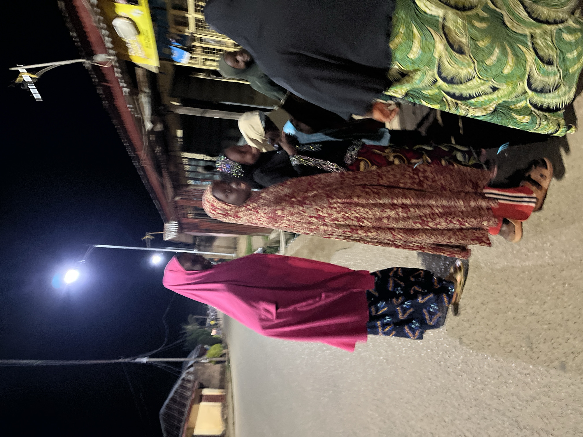 This is a nighttime street scene in Jos, Nigeria, illuminated by streetlights. Several people, primarily women, are visible standing and walking on what appears to be an asphalt road.
In the foreground, a woman wears a bright pink head covering and a dark blue skirt or dress with a light, repeating pattern. Behind her, another woman is dressed in a full-length, reddish-brown garment with intricate patterns, paired with sandals. Further back, several other individuals are present, some also wearing head coverings. One person is partially visible in a green and yellow patterned top, and another in a dark, abaya-like garment. A person in a green hoodie can be seen among the group.
The activity appears to be a casual gathering or passage of people on the street during the evening. The streetlights are a prominent feature, casting bright light onto the road and illuminating the immediate surroundings, contrasting with the dark night sky. Overhead wires or cables are visible stretching across the scene. Buildings, possibly shops or residences, line the street in the background, some faintly lit.
A notable detail is a yellow sign on a building in the upper left background, which has partially visible text including the word "SHOP" and possibly "FASHION." The overall atmosphere is calm, with people dressed in modest attire common to the region. The image is taken at night, indicated by the dark sky and reliance on artificial lighting.