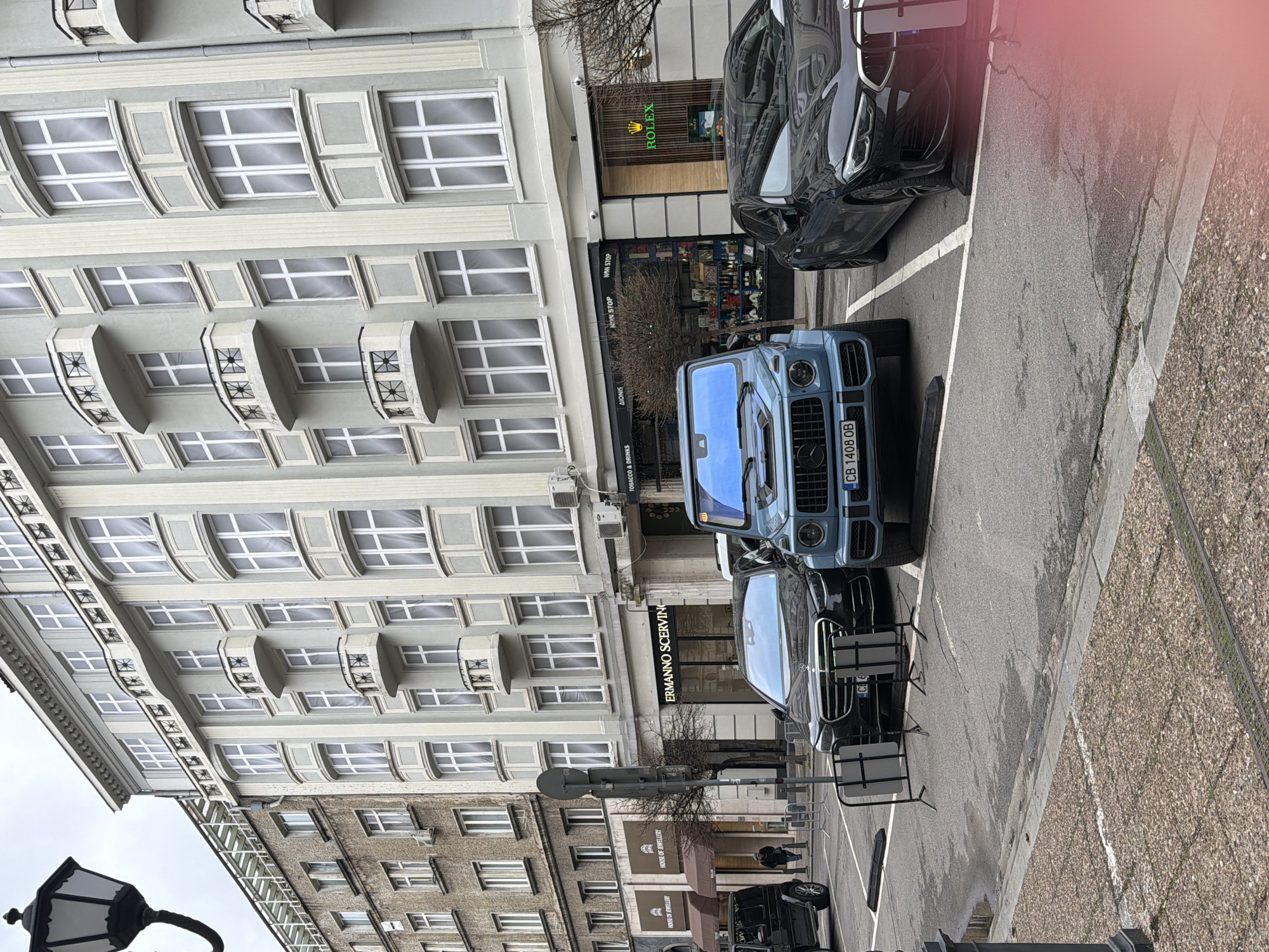 The image depicts a street scene in Sofia, Bulgaria, captured from a slightly elevated perspective. The focus is on a street lined with buildings, showcasing architectural details like windows and balconies. On the right side, parked cars, including a blue G-Wagon with license plate "CB 1408 OB", occupy the parking spaces.
The street level features shops like "HOUSE OF JEWELLERY", "ERMANNO SCERVIN", "AIONIS" and "ROLEX", along with signage indicating "TOBACCO & DRINKS" and "NON STOP". Street furniture such as a lamppost and crowd control barriers are visible.
The image presents a realistic portrayal of an urban environment, highlighting both the architectural and commercial aspects of the street.