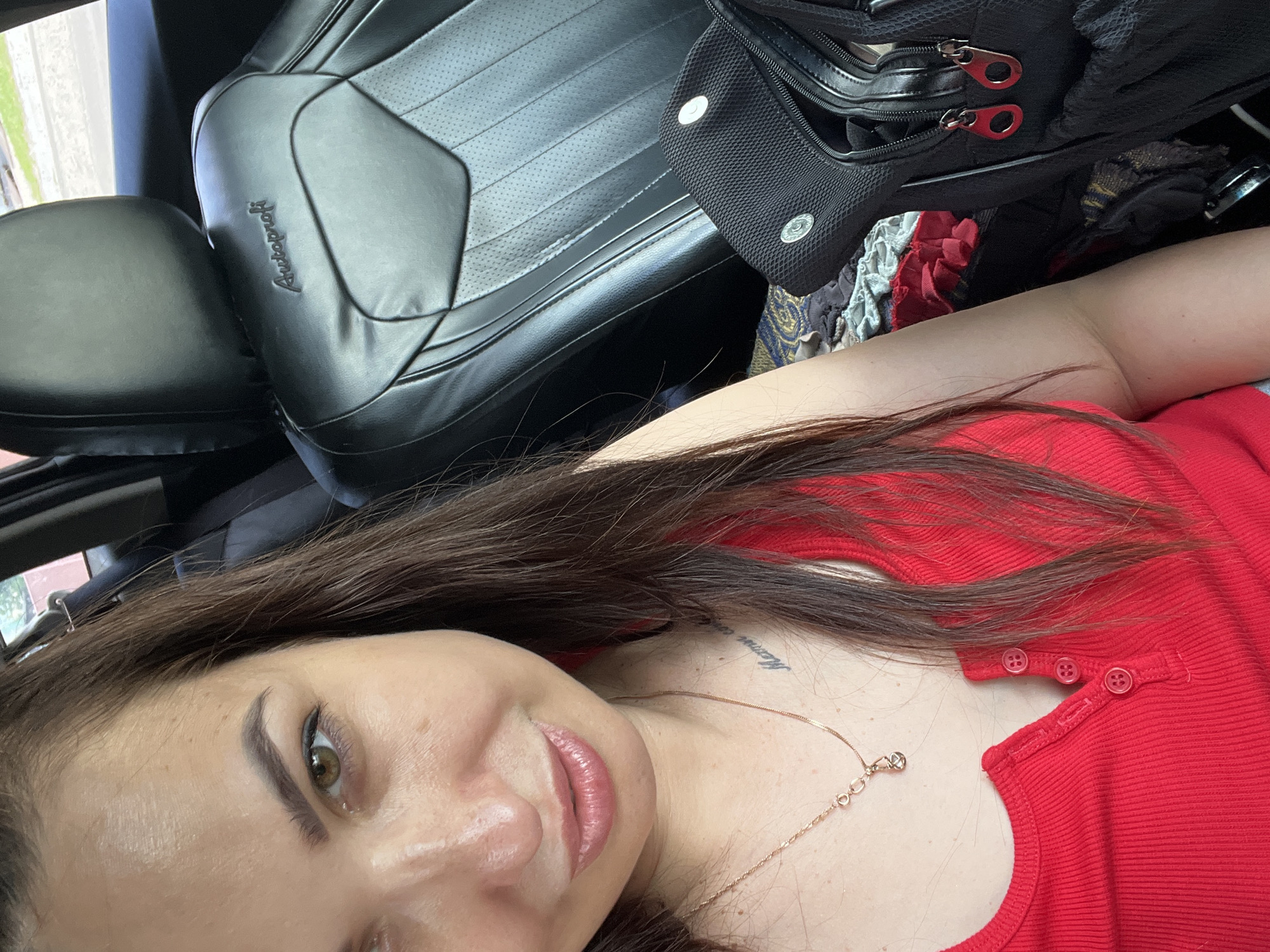 The media file is a close-up, angled selfie taken from a person's perspective inside a vehicle. The main subject is a woman with long, dark brown hair and fair skin, wearing a red top. She is looking towards the camera with her head tilted. Her makeup includes eyeliner and lipstick. A delicate gold necklace with a small charm is visible around her neck, resting on her collarbone. There is a faint tattoo on her neck.
In the background, the interior of a car is visible. A black leather car seat with a textured pattern and the embroidered word "Autoprofi" is prominently featured on the left side. A black bag with red zipper pulls is positioned above the car seat. Scattered fabrics with patterns are visible beneath the bag.
The image appears to be taken during the daytime, as suggested by the lighting. The setting is a car, likely parked or in transit, with a view of an outdoor scene with greenery visible through a window on the far left. No specific emotions are overtly displayed, but the selfie format suggests a personal moment. The only visible text is "Autoprofi" on the car seat.