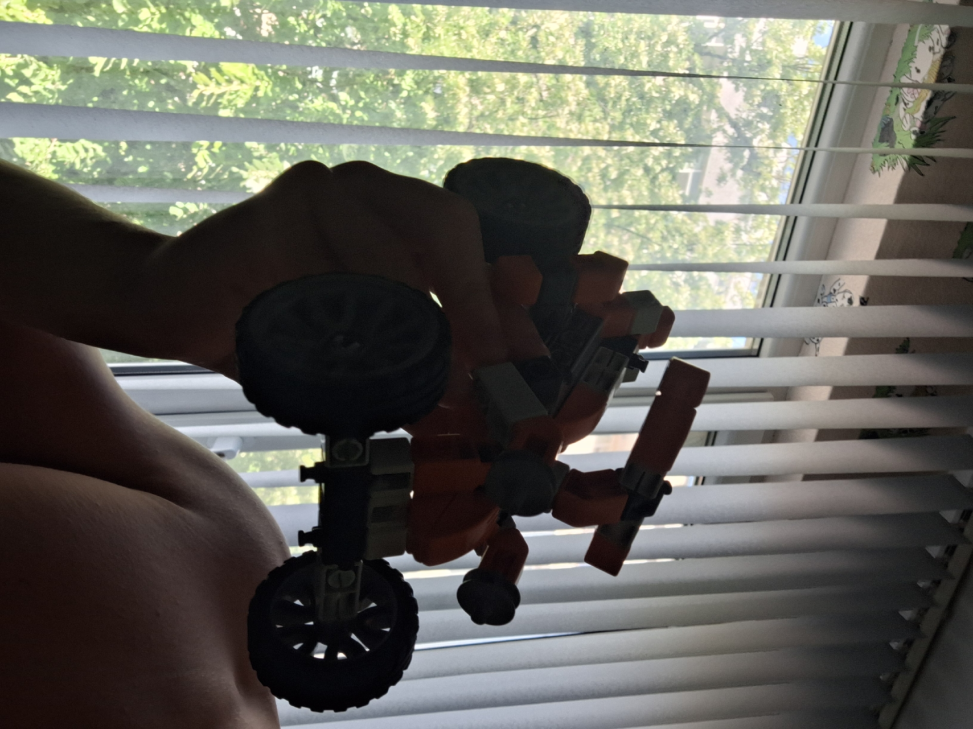 A close-up, silhouette-style shot shows a person's hand holding an orange and black Lego-style toy all-terrain vehicle in front of a window with vertical blinds. The background outside the window is blurry green foliage, suggesting an outdoor environment with trees. The person's shoulder and upper arm are visible in the lower right corner of the frame. The overall lighting suggests it is daytime, possibly late morning or early afternoon, with sunlight filtering through the blinds. The image does not provide any specific details that would allow for pinpointing a location within Varna, Bulgaria, or any other city.