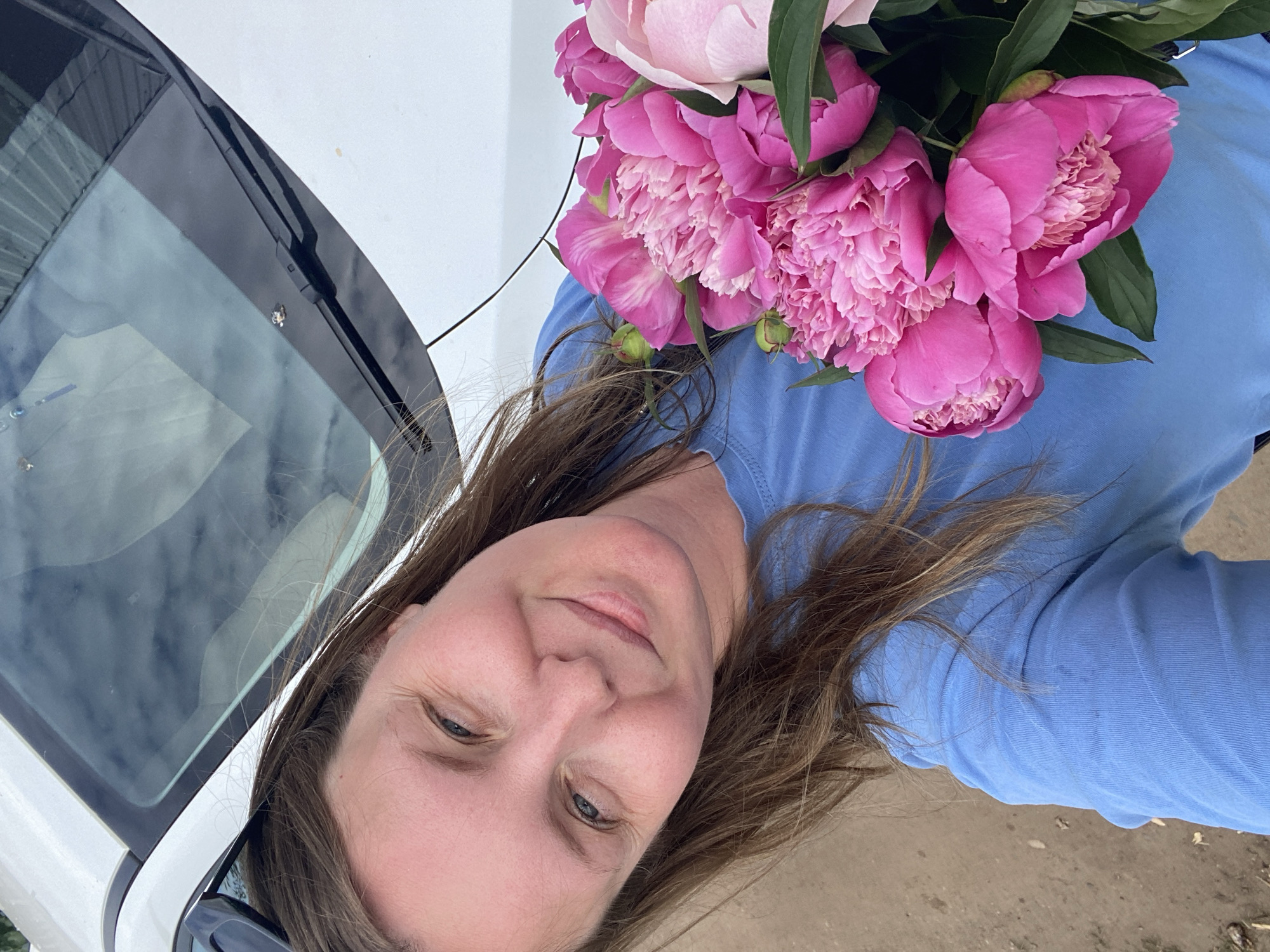 A medium shot captures a Caucasian woman with long brown hair and fair skin, tilted slightly to her left. She is smiling and looking towards the camera, her eyes open and her lips slightly parted. She is wearing a bright blue t-shirt. She is holding a large bouquet of pink peonies in her right arm, with several blooms prominently displayed. The bouquet features various shades of pink, from deep fuchsia to lighter blush tones. Green leaves and some unopened buds are also visible within the arrangement.
Behind the woman, a portion of a white car is visible, with its windshield and hood dominating the upper part of the frame. The windshield is partially reflecting the sky, which appears overcast with some lighter patches. A black windshield wiper is positioned on the right side of the windshield. The car's surface has a glossy sheen.
The lower portion of the image shows a ground surface that appears to be dirt or gravel, with some small debris scattered across it. The overall lighting suggests an outdoor setting, possibly during the daytime, with diffused light. The scene does not provide specific indicators of its precise location within the city of Chaikovsky, Russia, beyond it being an outdoor environment with a vehicle present.
