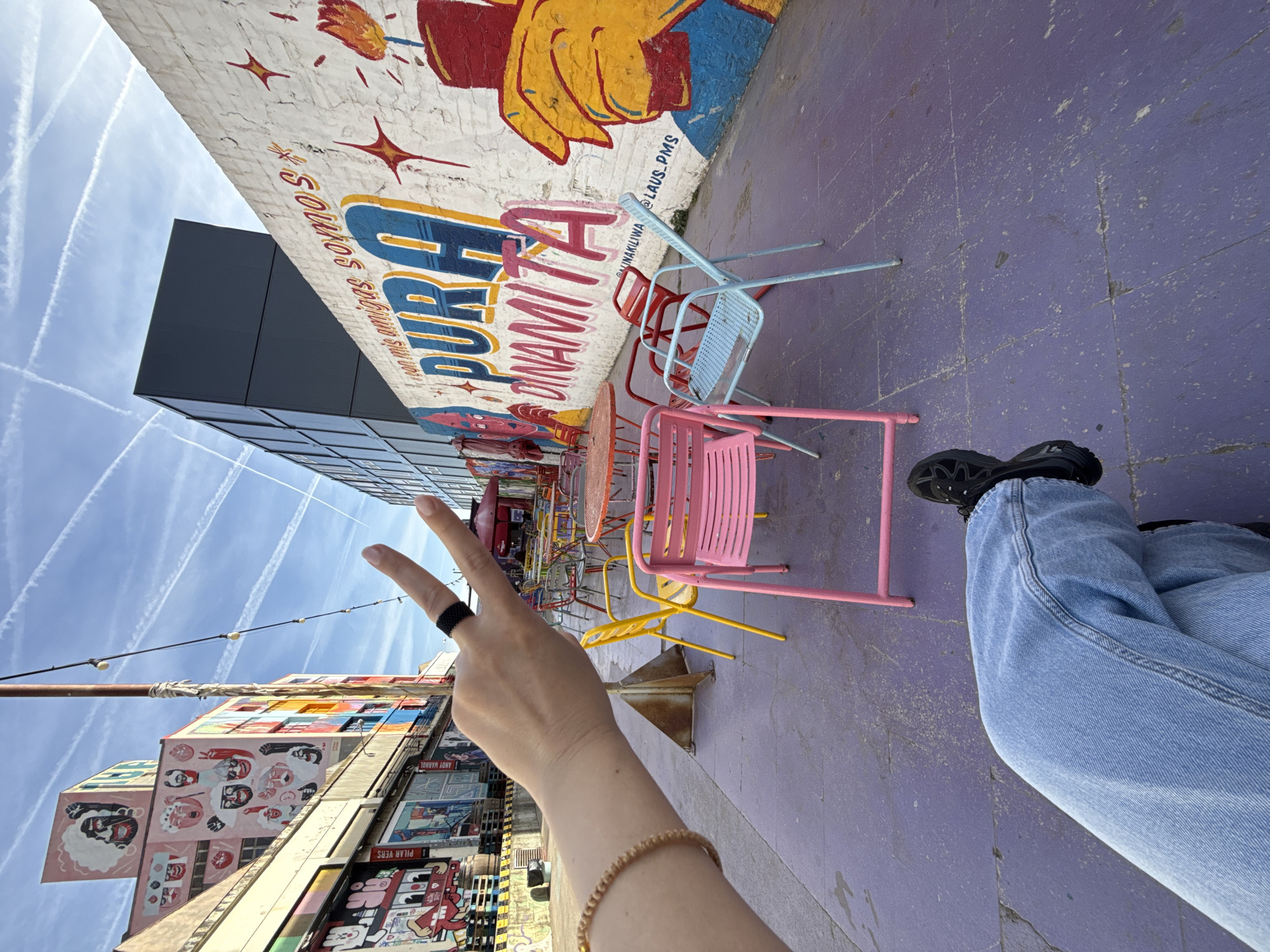 A ground-level, tilted outdoor shot depicts a person seated at a cafe in what appears to be Barcelona, Spain, identifiable by the artistic mural and architecture. The person's legs, clad in light blue jeans and black sneakers, are visible from the knees down. The person is giving a peace sign. The cafe has colorful chairs (pink, blue, red, yellow) arranged around tables on a purple-painted surface. A mural depicting a hand holding something on fire is visible. Across the street, buildings adorned with graphic art and portraits can be seen. The sky above is blue with contrails.