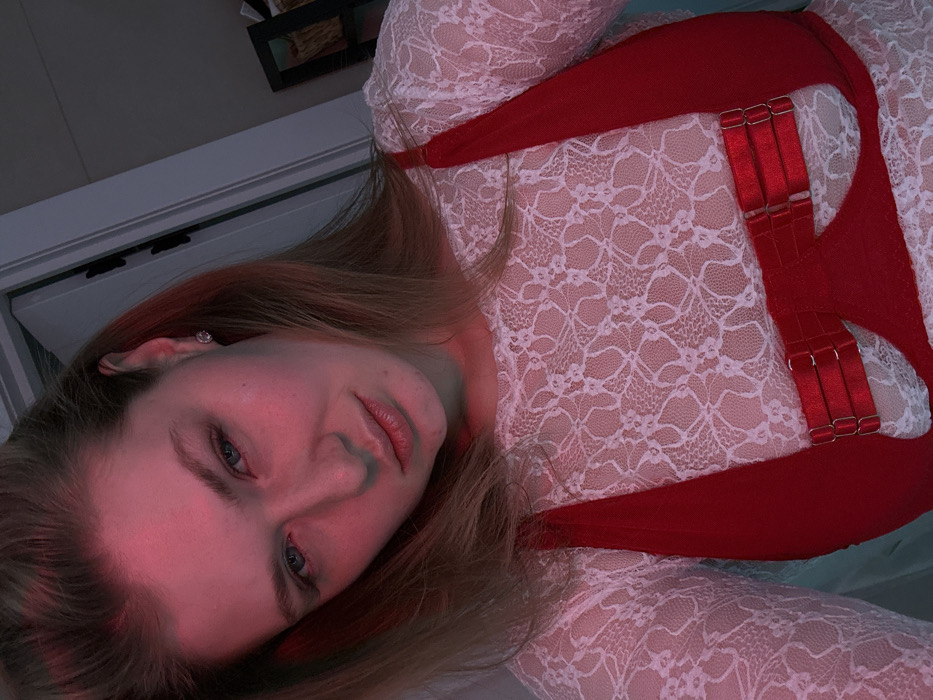 A woman with fair skin and light brown hair is positioned at an angle in the frame. She wears a white lace top paired with a red harness featuring adjustable straps with metal hardware. The light casts a red hue on her face, accentuating her features. Her gaze is directed toward the viewer. The background includes a partially visible door and shelf-like structures, contributing to a personal, indoor setting.