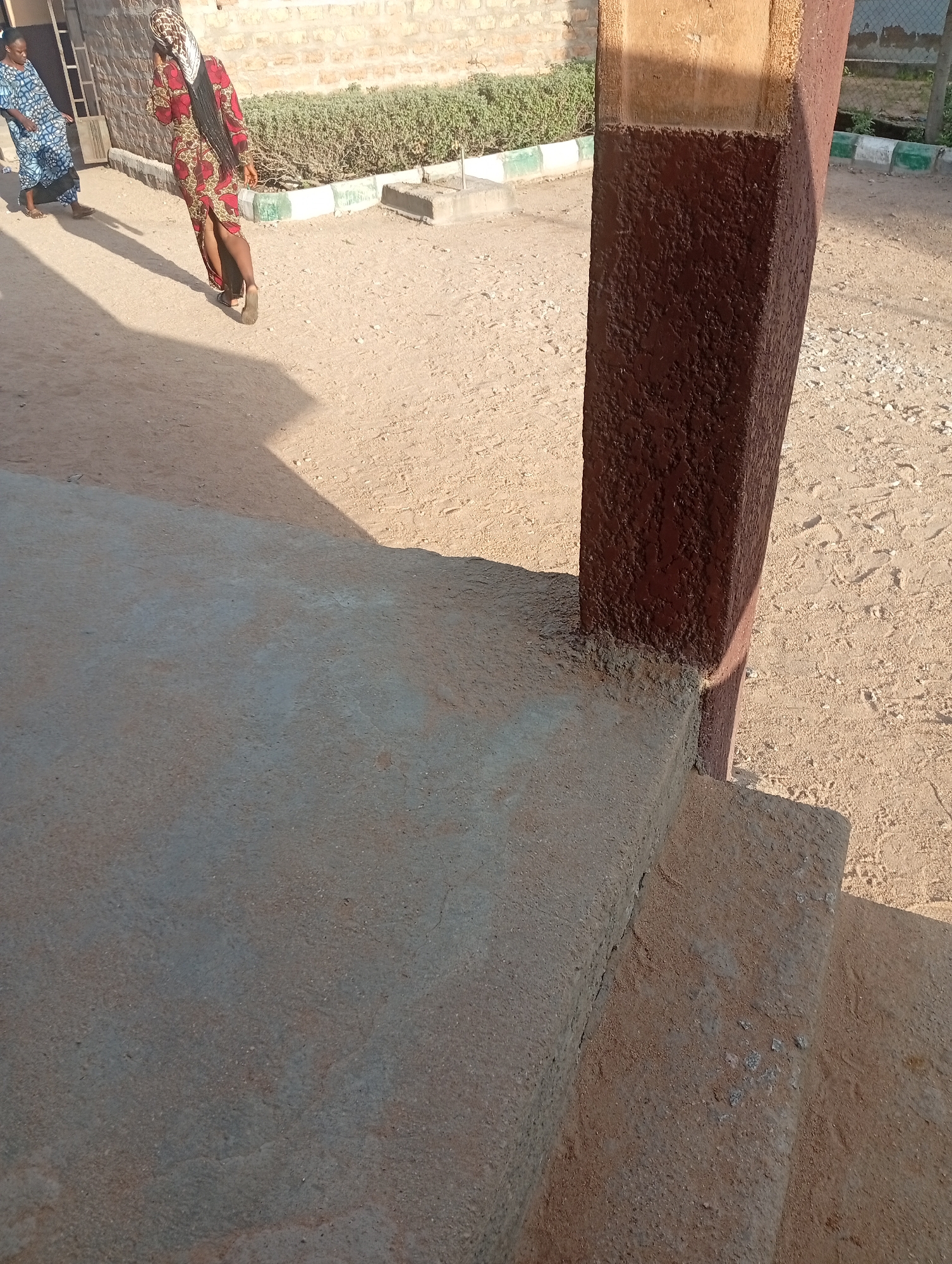 In Jalingo, Nigeria, two women are captured in a photograph. The woman in the foreground is walking away from the camera, her right leg slightly ahead of her left. She is wearing a red dress with a dark pattern, a headscarf, and appears to have dark hair. The second woman, in the background, is wearing a blue and white patterned dress and is walking to the left, away from the camera and further from the first woman. The environment consists of a dusty, sandy ground, with a concrete step in the foreground casting a shadow. In the background, there is a brick building, a hedge, and a fence. A large brown pillar stands to the right of the frame, partially obscuring the background and casting a shadow on the concrete step. The scene appears to be outdoors on a sunny day.