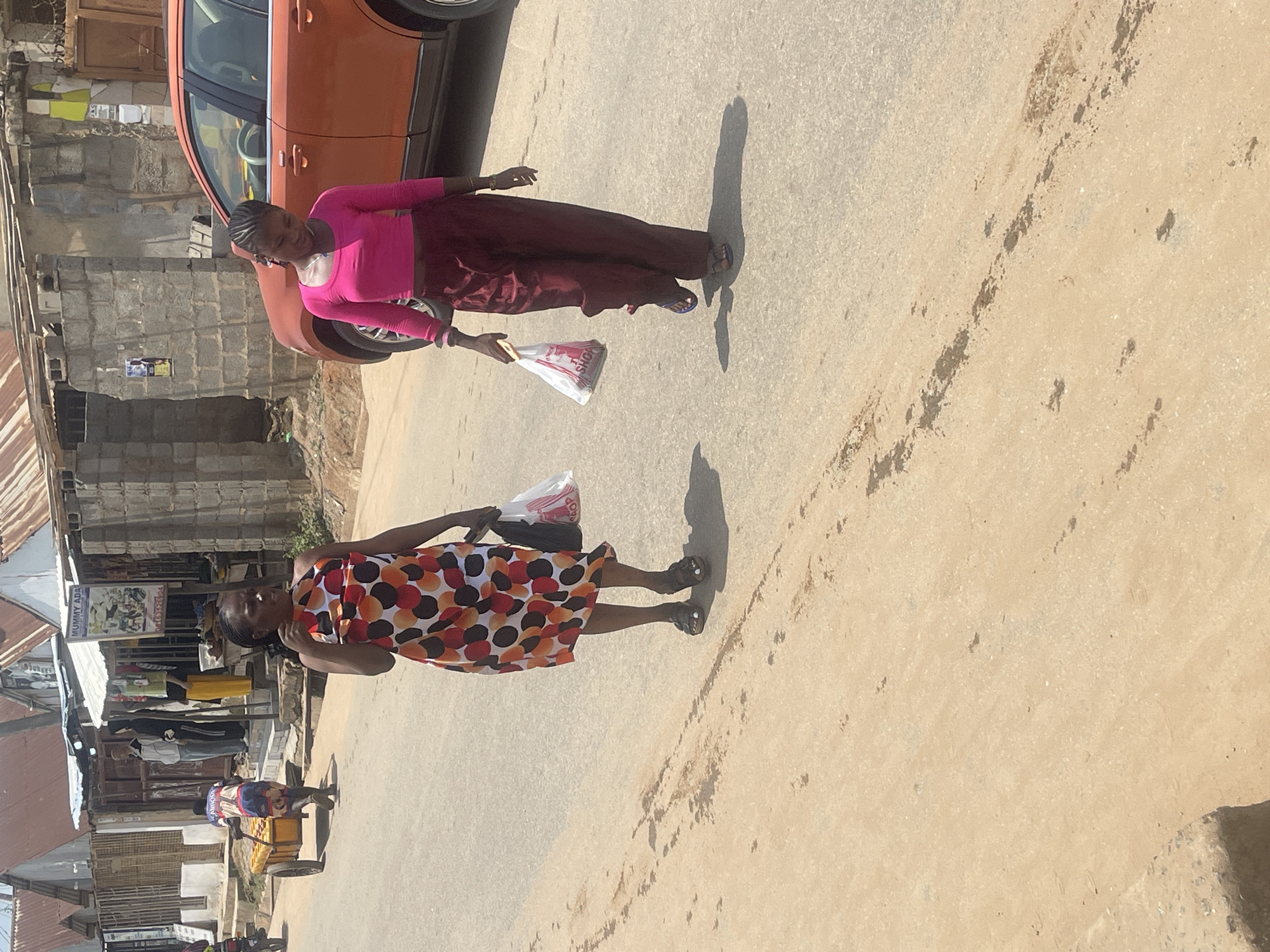 Two women are walking down a street in what appears to be a market area in Abuja, Nigeria. The scene is outdoors, with buildings and structures lining the street. The ground is paved with a sandy-colored material, and the lighting suggests it is daytime.
The woman in the foreground is wearing a dress with a colorful pattern of red, orange, and black circles. She is walking on the right side of the frame and appears to be carrying a white plastic bag in her left hand. Her hair is braided.
The woman in the background is wearing a bright pink long-sleeved top and a dark maroon skirt. She is walking on the left side of the frame, closer to an orange car. She is also carrying a white plastic bag in her right hand. Her hair is also braided.
Various stalls and shops are visible in the background, indicating a busy commercial area. One stall has a sign with text that appears to read "MUMMY ADA" and a phone number. The overall atmosphere seems to be that of daily life and commerce in a Nigerian city. The shadows suggest it is likely midday.