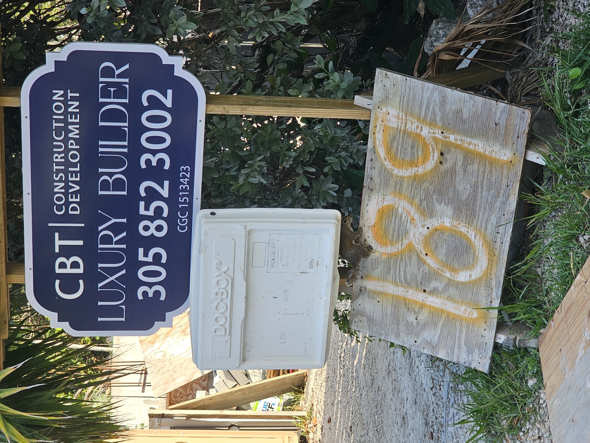 A low-angle outdoor shot captures a street sign in Islamorada, United States of America. The sign, a dark blue rectangle with rounded corners and a white border, reads "CBT CONSTRUCTION DEVELOPMENT LUXURY BUILDER 305 852 3002 CGC 1513423". Below the sign, a white "DOC-BOX" hangs from a wooden post, its face featuring spaces for "LOT", "JOB", "PERMIT", and "BLDC." A small squirrel is perched on a tilted wooden board in the foreground, which is spray-painted with the numbers "189" in yellow and white. The background consists of foliage, palm fronds, construction materials, and gravel.