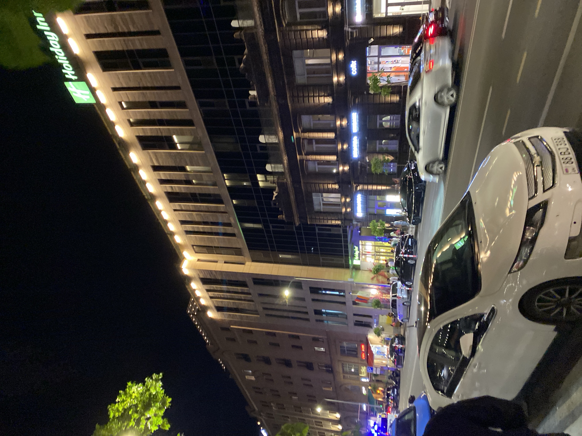 The image presents a vibrant nighttime street scene in Yerevan, Armenia, characterized by illuminated buildings and active traffic. The perspective is tilted, showcasing a bustling urban environment after dark.
On the left, a modern, multi-story building, identified by a prominent green "Holiday Inn" sign, stands brightly lit with strip lights along its facade. Adjacent to it, an older, more ornate building houses several businesses on its ground floor, marked by various illuminated signs, including a light blue neon sign that reads "coffi".
The street, running parallel to the buildings, is populated with cars. In the foreground, a white sedan is clearly visible, displaying an Armenian license plate with the number "99 GG 599". Further down the road, another white car shows its illuminated brake lights, suggesting it is either slowing down or stopped. Other vehicles and a few blurry figures, likely pedestrians, can be seen near the storefronts, indicating general city activity.
Artificial lighting from the building facades, shop signs, and vehicle lights bathes the scene in a warm glow, contrasting with the dark night sky. A brightly lit green tree is visible in the bottom left corner, adding a natural element to the otherwise concrete surroundings. The overall impression is one of a lively city evening, with ongoing movement and commercial activity.