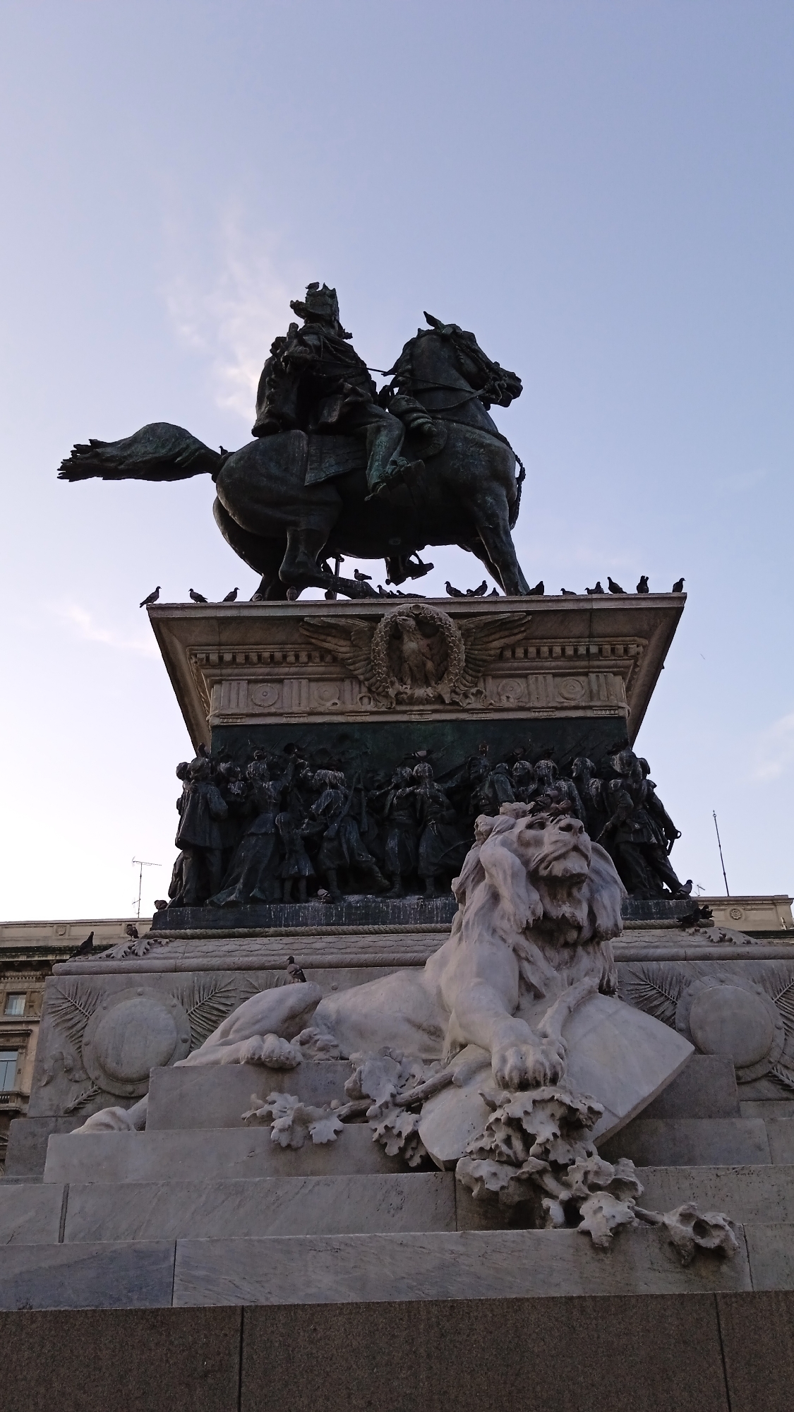 The image shows a monumental equestrian statue of Victor Emmanuel II, located in Piazza Duomo, Milan, Italy. The statue is made of bronze and depicts the king on horseback, wearing a crown and cape. The horse is rearing on its hind legs, with its front legs raised. The base of the statue is adorned with intricate reliefs and sculptures, including a marble lion lying at the front. Several pigeons are perched on the statue and its base, as well as on the surrounding architectural elements. The sky is clear and pale blue. The scene is captured from a low-angle perspective, emphasizing the grandeur and scale of the monument.