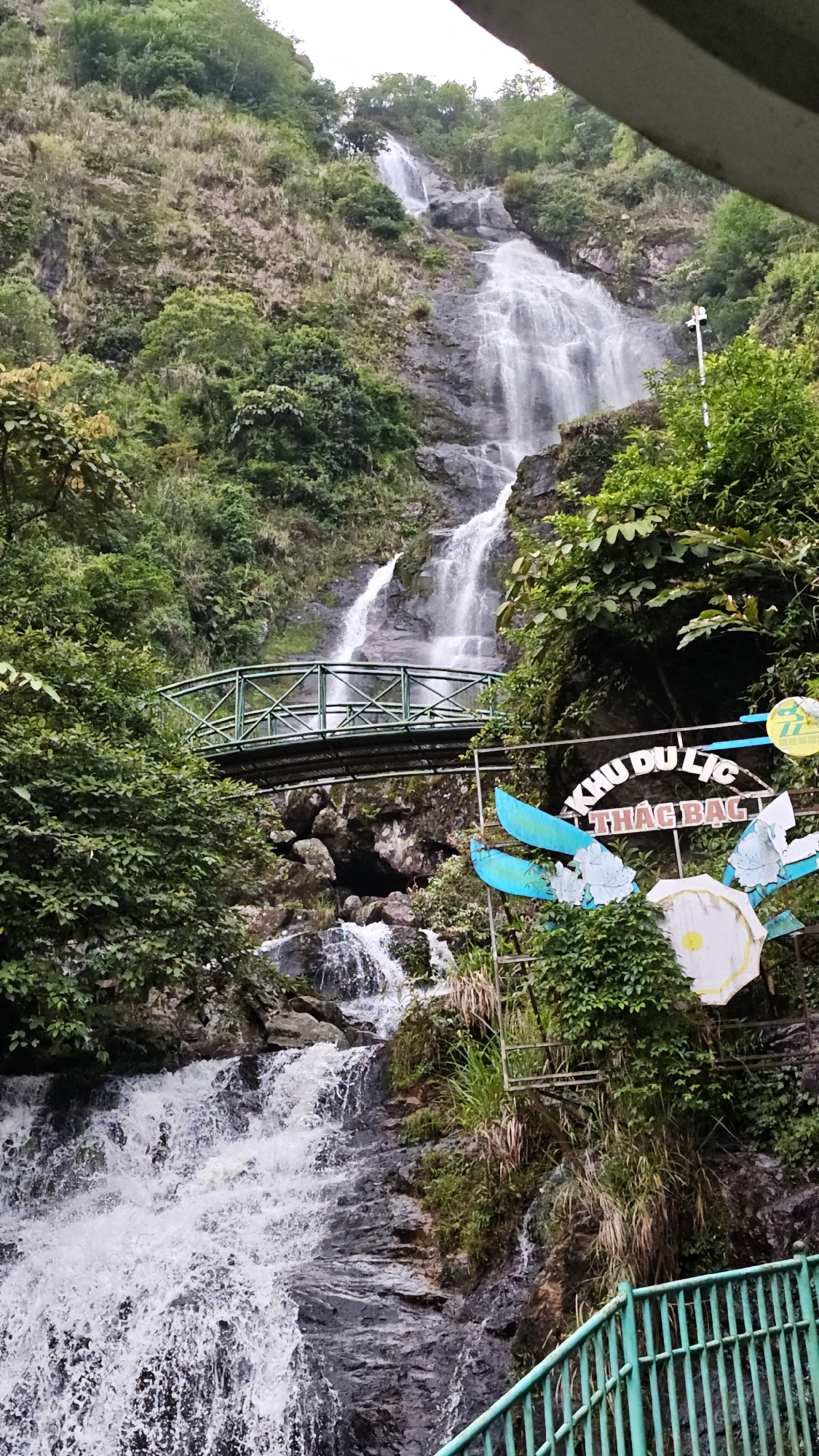 The image depicts a waterfall in Sa Pa Ward, Vietnam. The waterfall cascades down a rocky cliff face, surrounded by lush green vegetation. A green bridge spans a section of the waterfall. A sign with the words "KHU DU LỊCH THÁC BẠC" is visible near the bridge. The foreground includes a teal-colored railing.