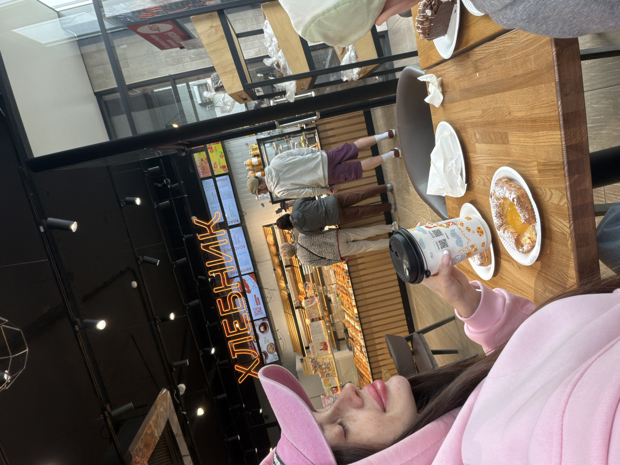 The image captures an interior view of a restaurant, likely in Saint Petersburg, Russia, as suggested by the Cyrillic text "ХЛЕБНИК" visible in neon sign lettering. The viewpoint is from a table, where a woman in a pink hoodie and hat is holding a disposable coffee cup with a patterned design, the focus of the foreground. On the wooden table are two white plates, one with a pastry filled with jam and another that is empty.
Several individuals are visible in the background. There is a queue of patrons waiting to be served at a bakery display counter. The environment appears to be a well-lit cafe setting with wooden shelves and a glass display case showcasing various baked goods. The restaurant is near a window.