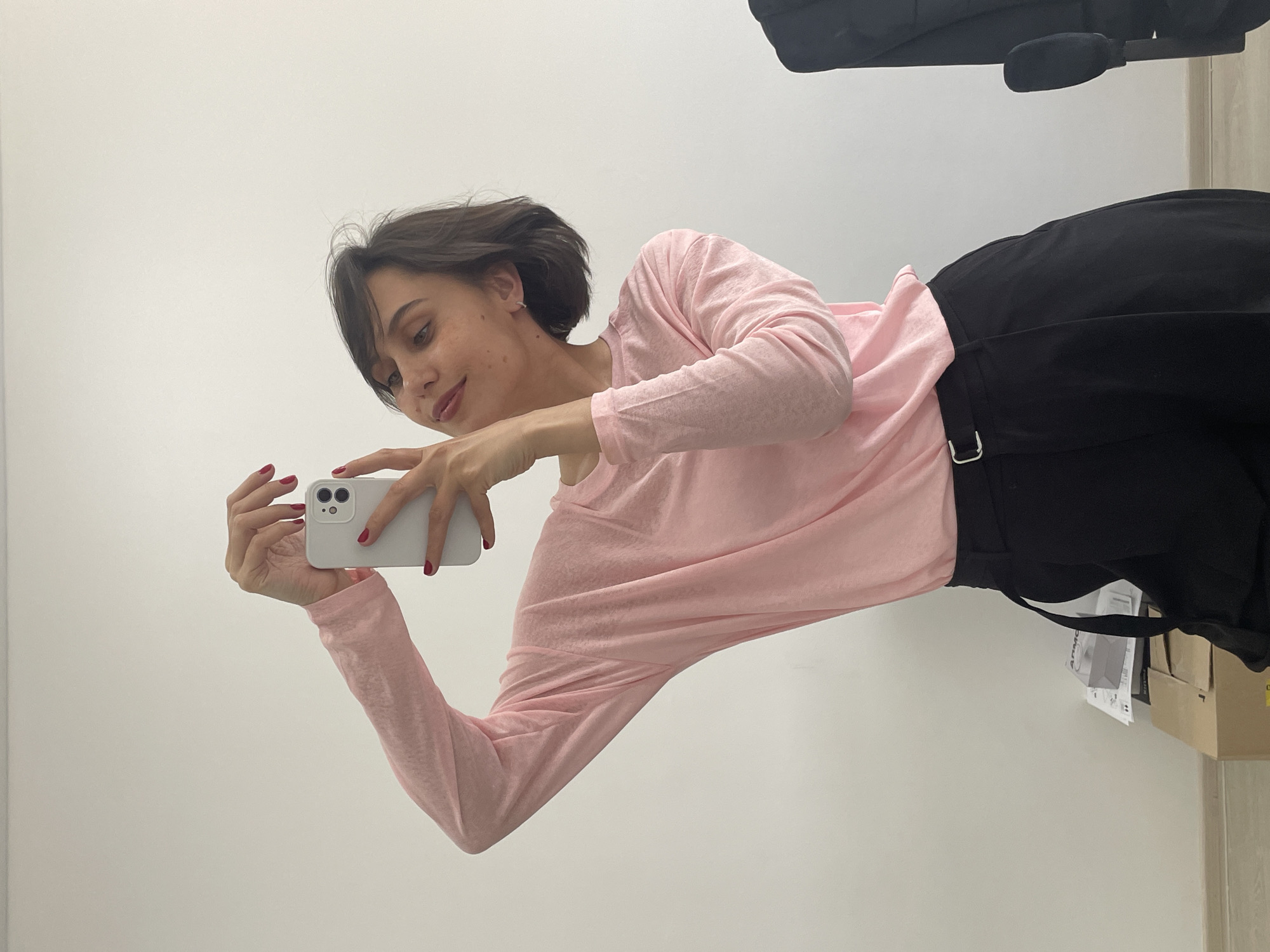 A woman is standing in front of a white wall and holding a white smartphone to take a picture of herself. She is wearing a long-sleeved pink shirt and black pants. Her hair is short and dark. The location is a room with a white wall and cardboard boxes on the floor. There is a chair visible in the background. There is no indication that this image was taken in Izhevsk, Russia.