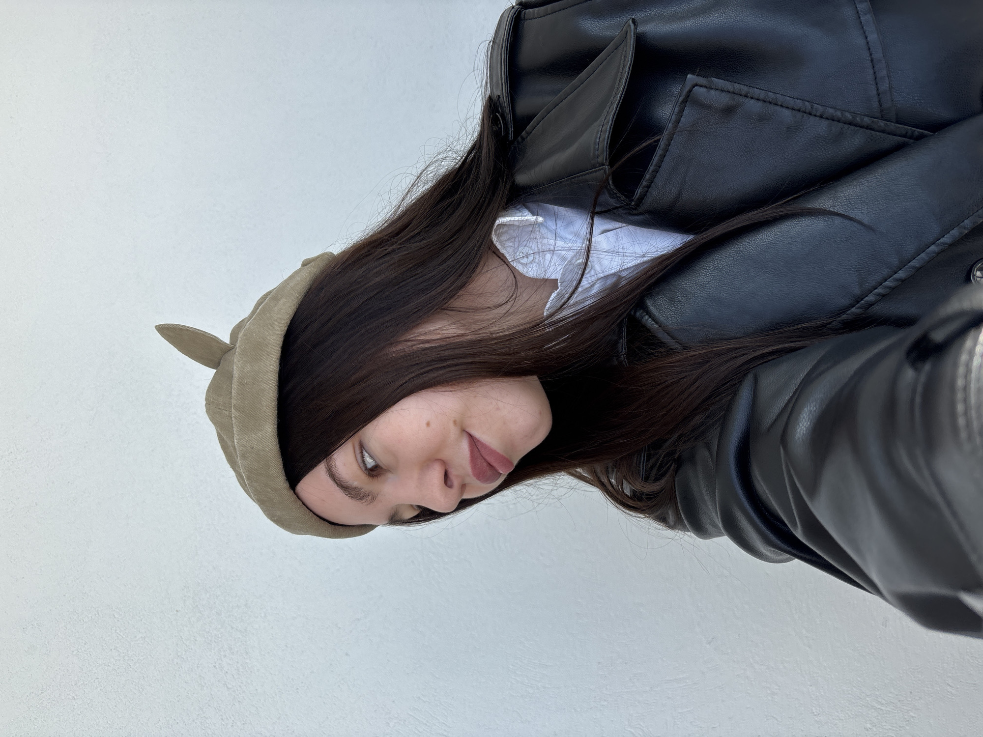 The scene is located in Perm, Russia. A single female subject is depicted in a chest-up, close-up portrait against a textured white wall. She wears a light olive-green or beige cap featuring an upward-projecting detail on the right side, a white collared shirt with ruffled details, and a black leather or faux leather trench-style jacket. Her long, dark brown hair is styled straight and falls over her shoulders. The subject's head is slightly tilted, and her gaze is directed upwards and to her right, away from the camera's lens. Her right eye is partially visible, and her lips are tinted with a reddish-pink color, slightly parted. Multiple small moles are visible on her right cheek. The lighting is bright and even.