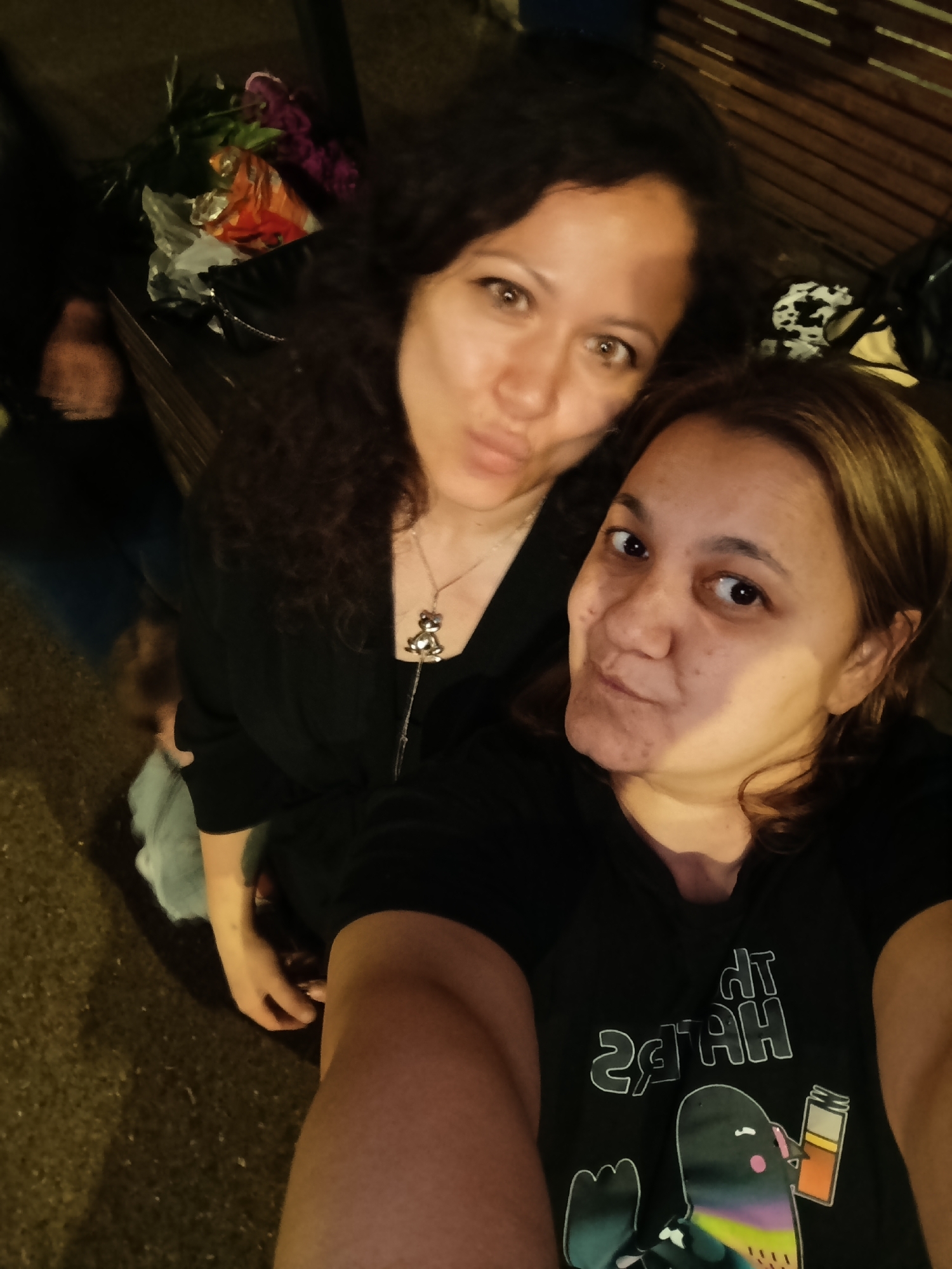 The image shows a selfie of two adult females, taken from a high angle. The woman in the foreground, on the right, has medium-length brown hair and is wearing a black t-shirt with a graphic of a cartoon bird and text. The woman in the background, on the left, has long, dark, curly hair and is wearing a black top with a necklace. Both women are looking at the camera and appear to be posing. The background includes a wooden slatted surface, some foliage, and what appears to be a person in the lower left portion of the image, possibly a child. The scene suggests an outdoor setting at night, with the lighting casting shadows. There is no discernible information in the image that would indicate a specific location within the city of Chaikovsky, Russia.