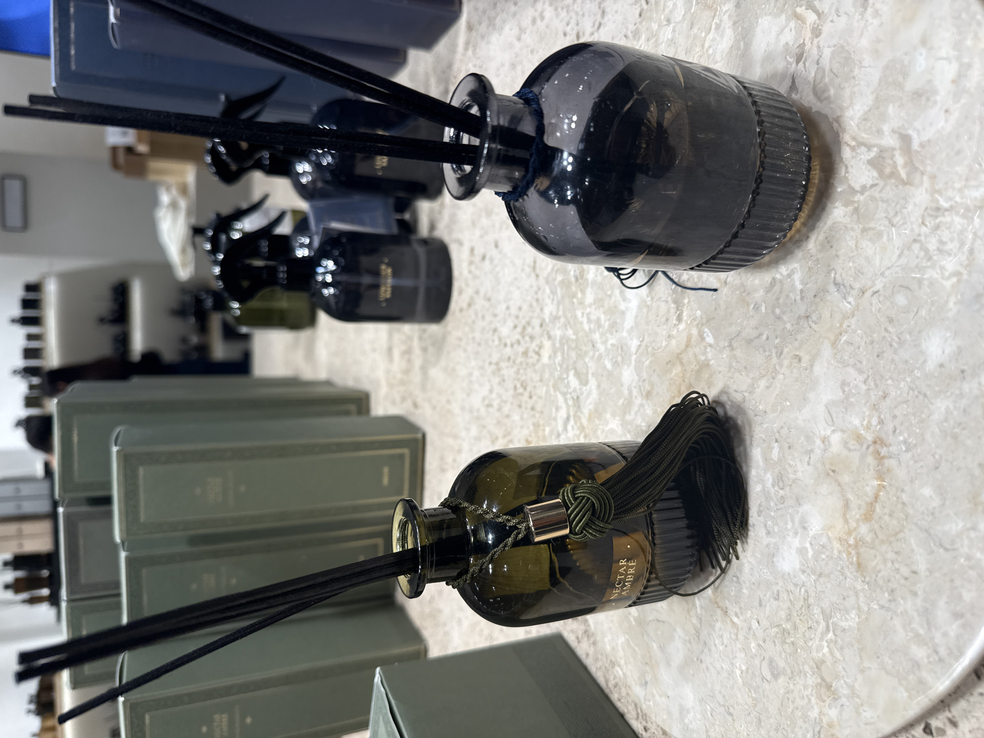 In Almaty, Kazakhstan, an indoor retail scene unfolds, focusing on decorative home fragrance products. Two reed diffusers stand prominently on a marble countertop. The diffuser on the left, made of dark green glass, contains a golden liquid and is adorned with a dark green tassel and gold accents. It displays the label "NECTAR AMBRE." The diffuser on the right is made of smoky dark gray glass and has a dark blue cord around its neck. Both diffusers have black reeds inserted into their openings. In the background, several tall, slender, olive-green boxes are neatly stacked, some with embossed text. Further back, a white shelf displays various dark-colored bottles and spray dispensers, some appearing to be cosmetic or home cleaning products. The overall environment suggests a boutique or department store display. No individuals are clearly visible in the foreground or immediate background.