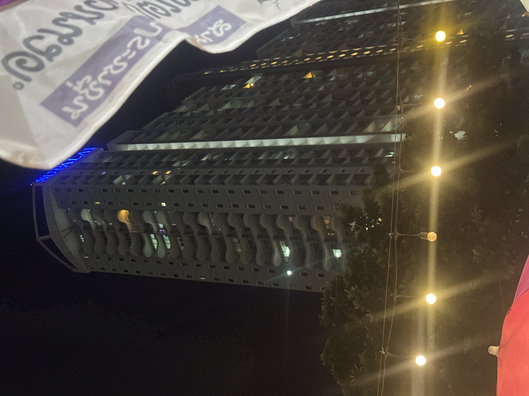 The image captures a nighttime urban scene, featuring a tall building, decorative lights, and a white banner with text. The perspective is from the ground looking up, with the image rotated 90 degrees clockwise from its natural orientation.
The main subject is a modern, high-rise building, likely a condominium or office block. Its facade features a repeating pattern of recessed windows or balconies, giving it a textured, grid-like appearance. Towards the lower floors, there's a distinctive architectural element: a series of wavy, organic-shaped balcony railings or decorative panels. Many windows on the building show internal lights, indicating occupancy. At the bottom left of the building, blue illuminated text clearly spells out "SUPPALAI".
On the right side of the frame, a vertical string of warm yellow string lights adds a festive or decorative touch, casting bright glows and starburst effects. Trees and foliage are visible around and behind these lights, suggesting some greenery in the urban setting.
Across the top of the image, a white banner is partially visible, featuring Thai script in purple and dark gray. While not entirely clear, some characters resemble "สุขสันต์" (Suk-san), which means "happy" or "joyful" in Thai, suggesting a celebratory message.
The scene is set at night, with the sky appearing dark. The artificial illumination from the building, street lights, and banner creates a vibrant, urban atmosphere. No specific human activity is evident. Given the visible Thai text and the location context, the scene is likely from Pattaya City, Thailand. A glimpse of a red or pink object, possibly another banner or decorative element, is visible on the far right edge.