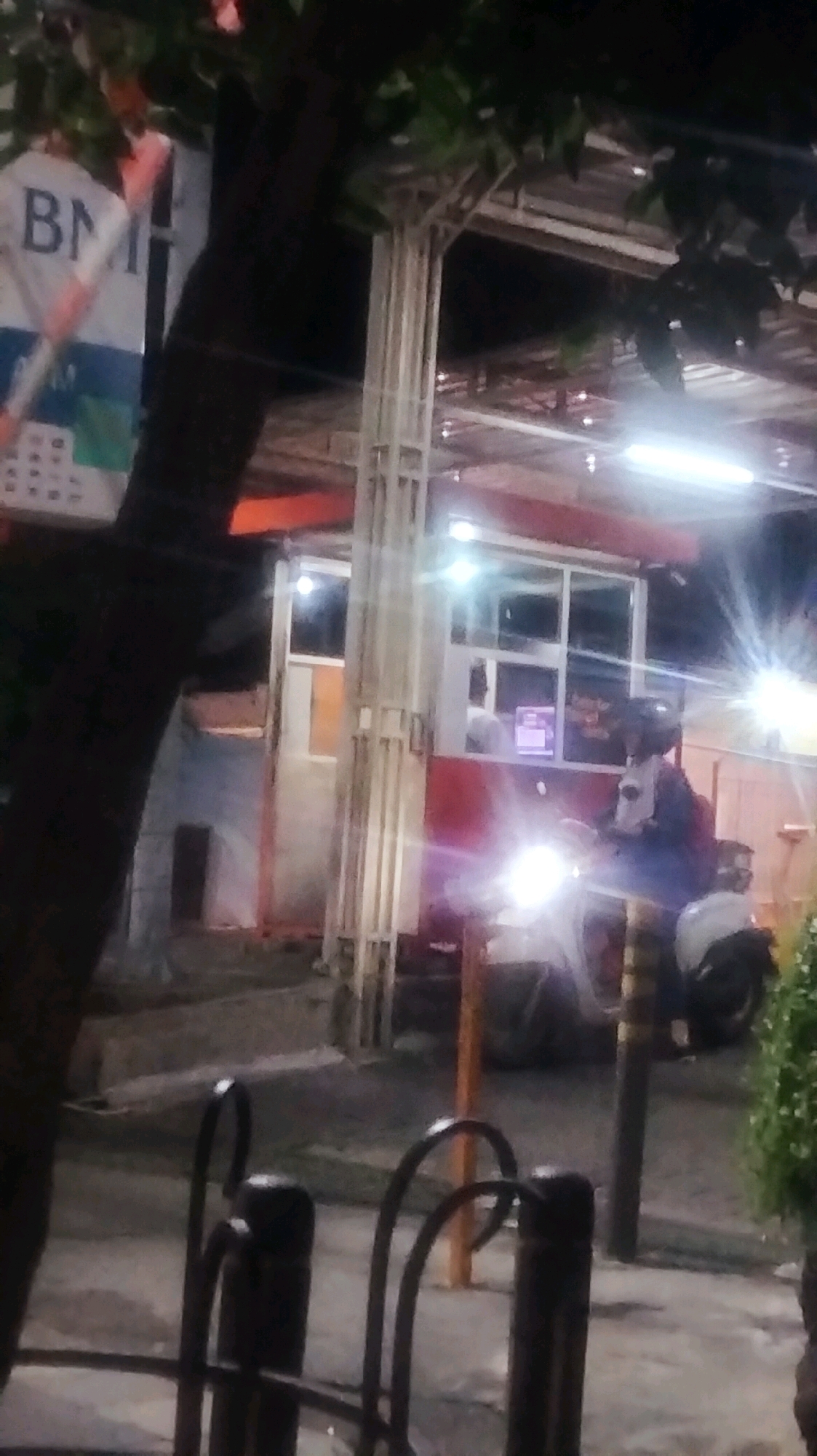 The image depicts a night scene in Bojonegoro, Indonesia, likely at a roadside food stall or service window. A dark tree trunk dominates the left foreground, partially obscuring a white sign with blue lettering that appears to read "BM". To the right, a structure with a red and white facade features large windows. A person wearing a helmet is seated on a motorcycle in front of the window, their face illuminated by the motorcycle's bright headlight. Another person is visible inside the structure behind the window. The ground is paved, and a metal railing or barrier is present in the lower foreground. The overall atmosphere is dimly lit, with artificial lights providing illumination.