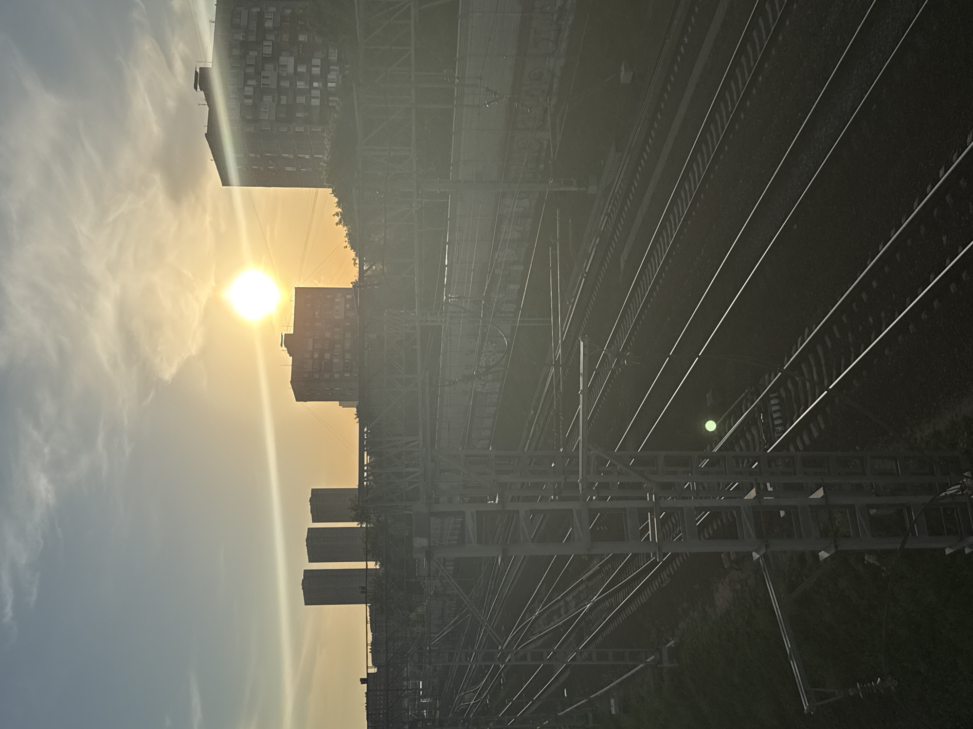 The image depicts a cityscape in Moscow, Russia, at sunset. The sun, low on the horizon, shines brightly behind buildings on the left. Silhouetted structures include a complex steel overpass, buildings of various heights, and overhead electrical lines. To the right of the buildings are several parallel railroad tracks. Vegetation is visible at the bottom and along the overhead electrical lines. The sky fades from yellow to blue.