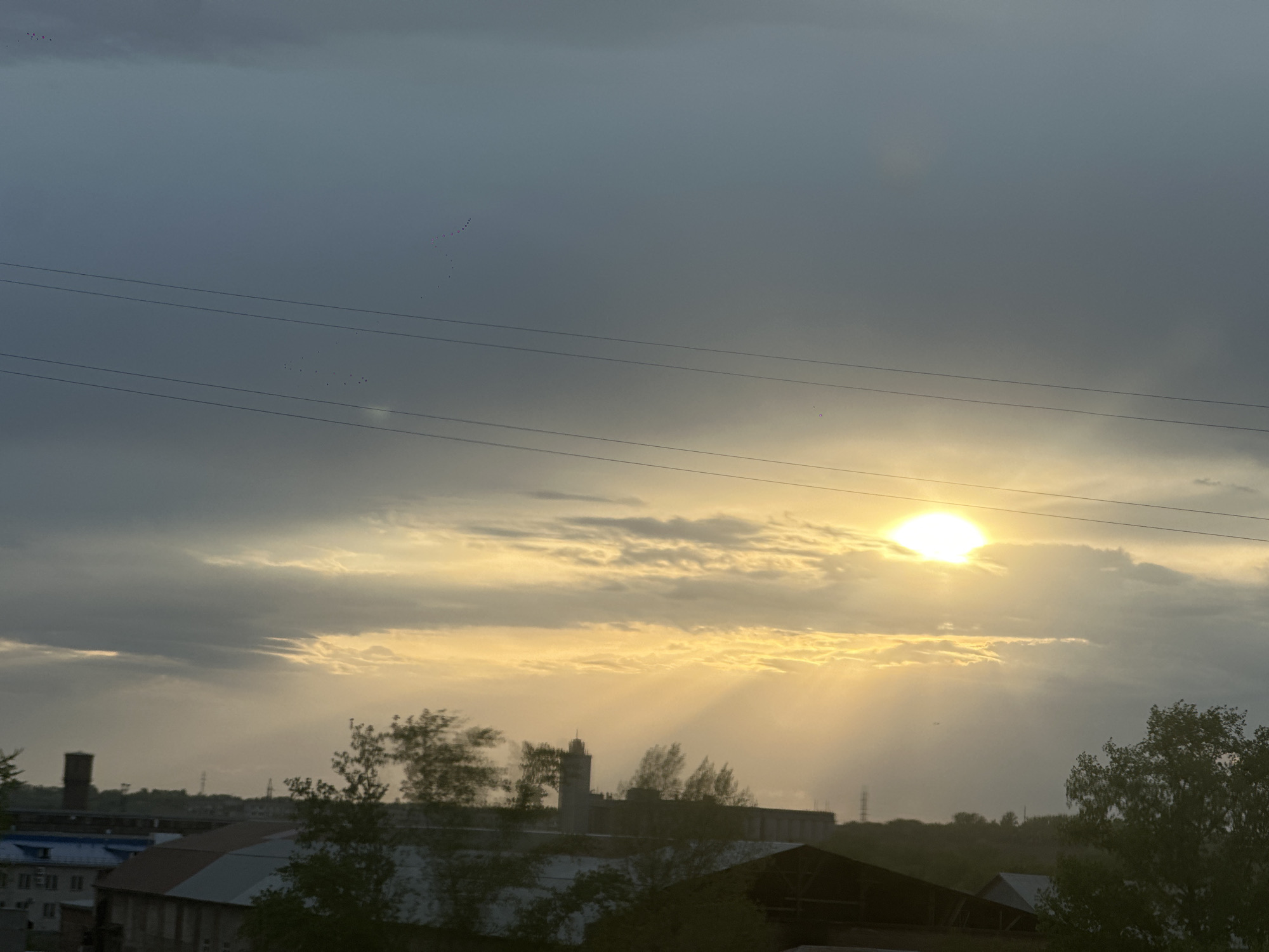 The image shows a view of a cloudy sky with the sun partially obscured, creating rays of light. In the foreground, there are rooftops and trees. In the mid-ground, there are industrial-style buildings with a tall structure that could be a tower or chimney. Overhead, there are power lines. The image was taken in Biysk, Russia. There are birds in the upper left quadrant, flying.