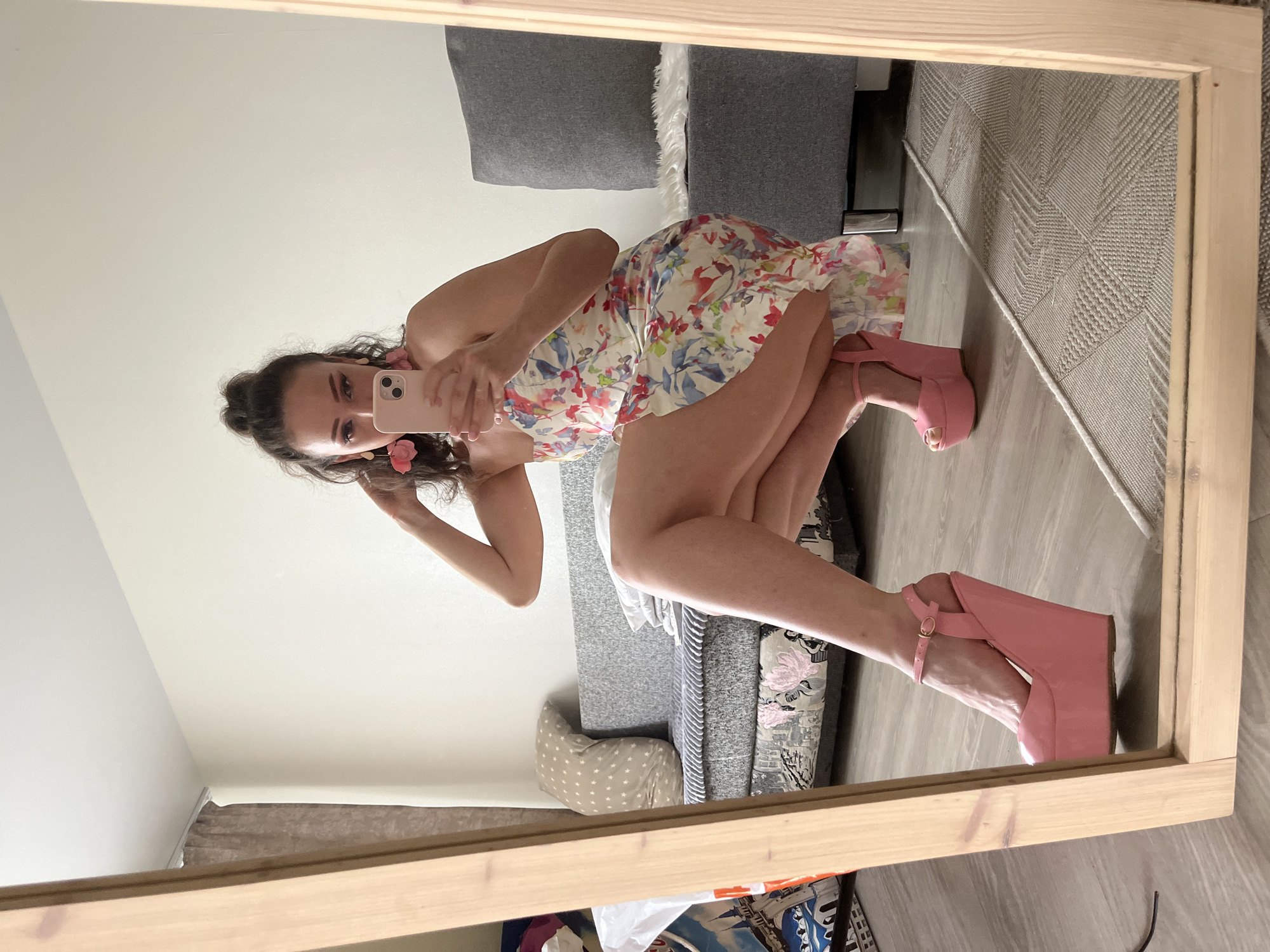 A woman is captured in a mirror's reflection, squatting and holding a smartphone to take a picture. She is wearing a floral print dress and pink wedge platform heels. The scene appears to be indoors, possibly a bedroom or living room, with furniture such as a sofa and pillows visible in the background. The flooring is wooden, and a rug is partially visible. There is no indication of the location being in the city of Kazan, Russia.