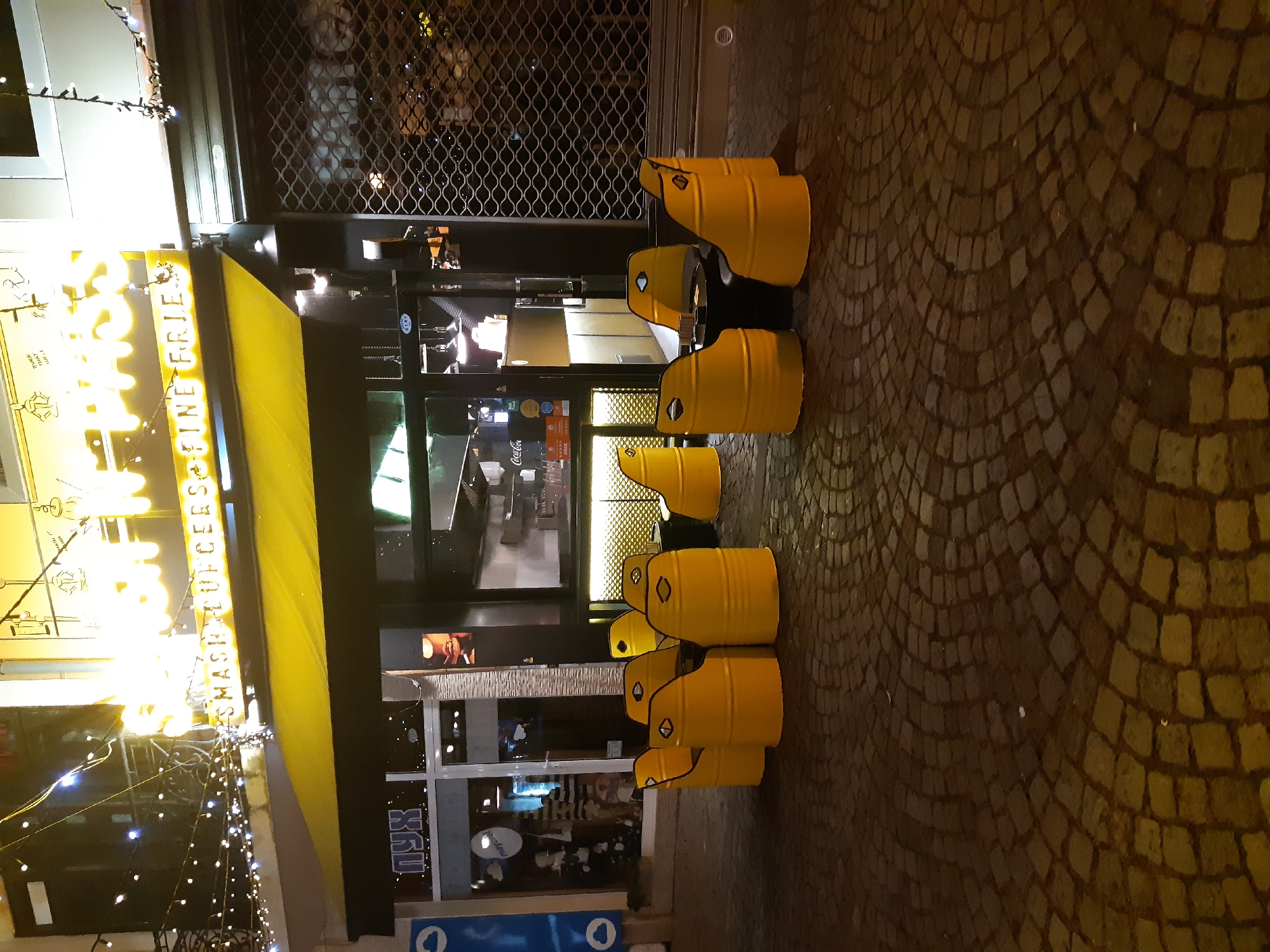 The image shows a street scene at night in Plovdiv, Bulgaria. The scene features a row of yellow oil drum chairs outside a restaurant named "SMASH BURCEES & FINE FRIES". The storefront has a yellow awning. There is decorative string lighting hanging from the awning. The restaurant's interior is visible through the glass windows, with what appears to be people inside. The street is paved with cobblestones.