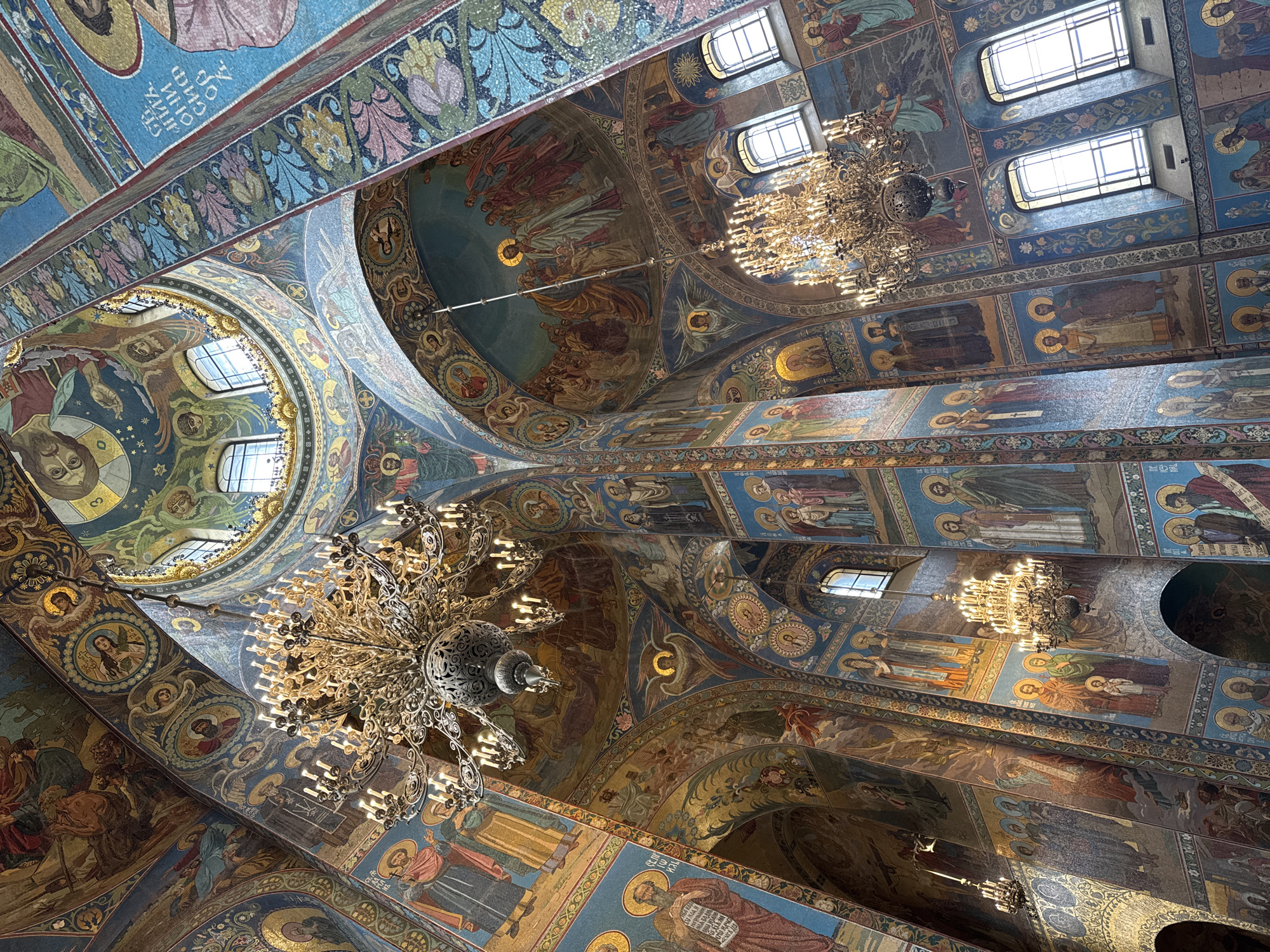The image captures the resplendent, mosaic-covered interior of a grand church or cathedral, looking upwards from below. Given the context, this is likely a prominent religious edifice in Saint Petersburg, Russia. Every visible architectural surface, including the vast domes, soaring arches, vaults, and supportive beams, is meticulously adorned with vibrant, intricate mosaic artwork.
The primary subjects depicted in the mosaics are numerous religious figures, such as saints, apostles, and biblical characters, rendered in traditional Orthodox iconographic style, often with golden halos. Large narrative scenes from biblical stories or the lives of saints are intricately portrayed across expansive panels. These mosaics utilize a rich and varied palette, prominently featuring blues, greens, golds, browns, and reds. Interspersed with the figural representations are elaborate geometric and floral patterns that frame the scenes and embellish the architectural elements.
Several multi-tiered, ornate chandeliers, crafted from what appears to be metal and fitted with numerous light bulbs resembling candles, hang from the ceiling. These chandeliers are illuminated, contributing a warm glow that complements the natural light filtering in through several high-set arched and rectangular windows.
While no direct activity is taking place in the image, the elaborate religious artistry signifies a profound space of worship and spiritual contemplation. Visible Cyrillic text, such as "СИНА БОЖIЯ" (Son of God), can be discerned on some mosaic-covered beams, identifying figures or conveying sacred inscriptions common in Orthodox architecture. The interplay of natural light and the illuminated chandeliers suggests a daytime setting within this immense and ornately decorated structure.