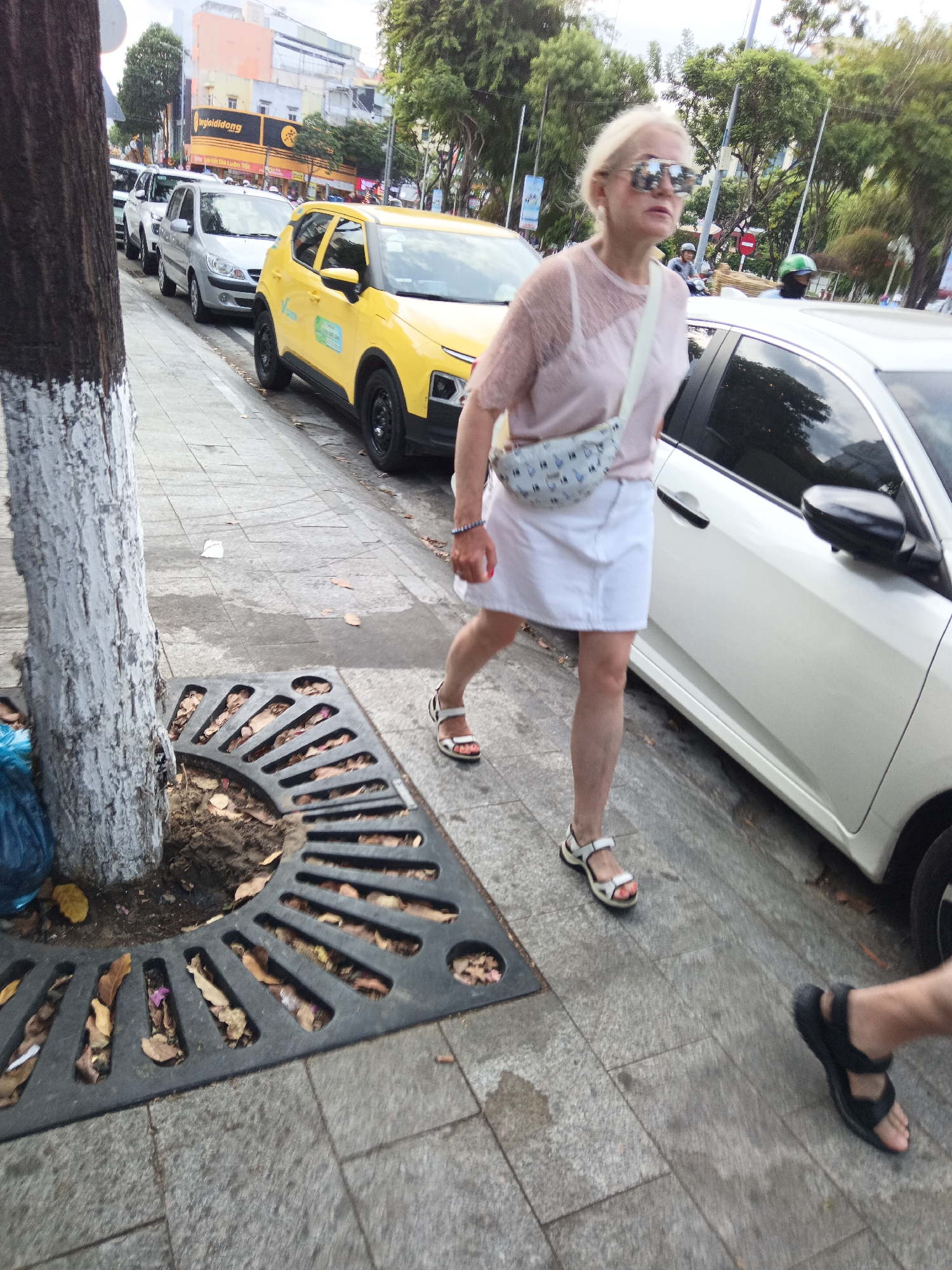 In Nha Trang, Vietnam, a blonde woman wearing a light pink lace top, a white skirt, white sandals, and sunglasses walks along a sidewalk next to a line of parked cars, including a yellow taxi. She carries a light-colored crossbody bag with a repeating bird pattern. Another person's legs, wearing black sandals, are visible in the foreground. The sidewalk is paved with gray tiles, and a tree with a white-painted trunk stands to the left, next to a black metal tree grate filled with fallen leaves. Buildings and trees are visible in the background.