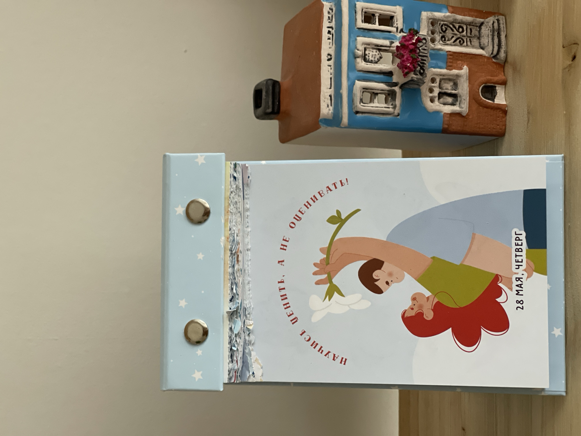 A light blue flip calendar with white stars, featuring a cartoon illustration of two people holding a flower branch, sits on a wooden surface. Text in Cyrillic script is visible on the calendar page. Next to the calendar is a miniature, brown-and-blue painted house, depicting a traditional European building with flower boxes on the windows. The house is likely a decorative item or souvenir. The background is a beige wall. The scene gives the impression of a domestic setting, possibly a shelf or desk arrangement. The image does not contain any information that explicitly identifies the location as Izhevsk, Russia.