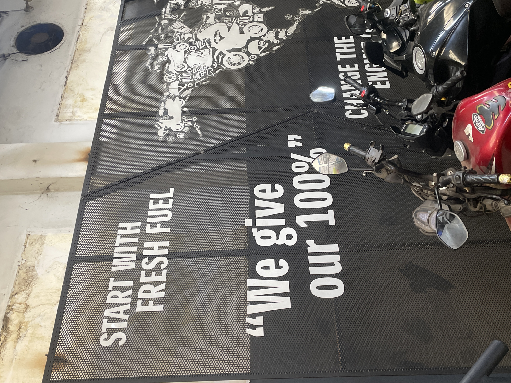 This indoor shot shows a section of a perforated metal wall display featuring white lettering against a black background. The words "START WITH FRESH FUEL" are visible on one panel, while the phrase ""We give our 100%"" appears on another panel. The top panel displays a motorcycle engine schematic in white. Part of a motorcycle's handlebars and rearview mirror are visible in the foreground, overlapping the display. The wall behind the display is weathered and stained, with a circular ventilation opening near the top left corner. The image was taken in Dhaka, Bangladesh.