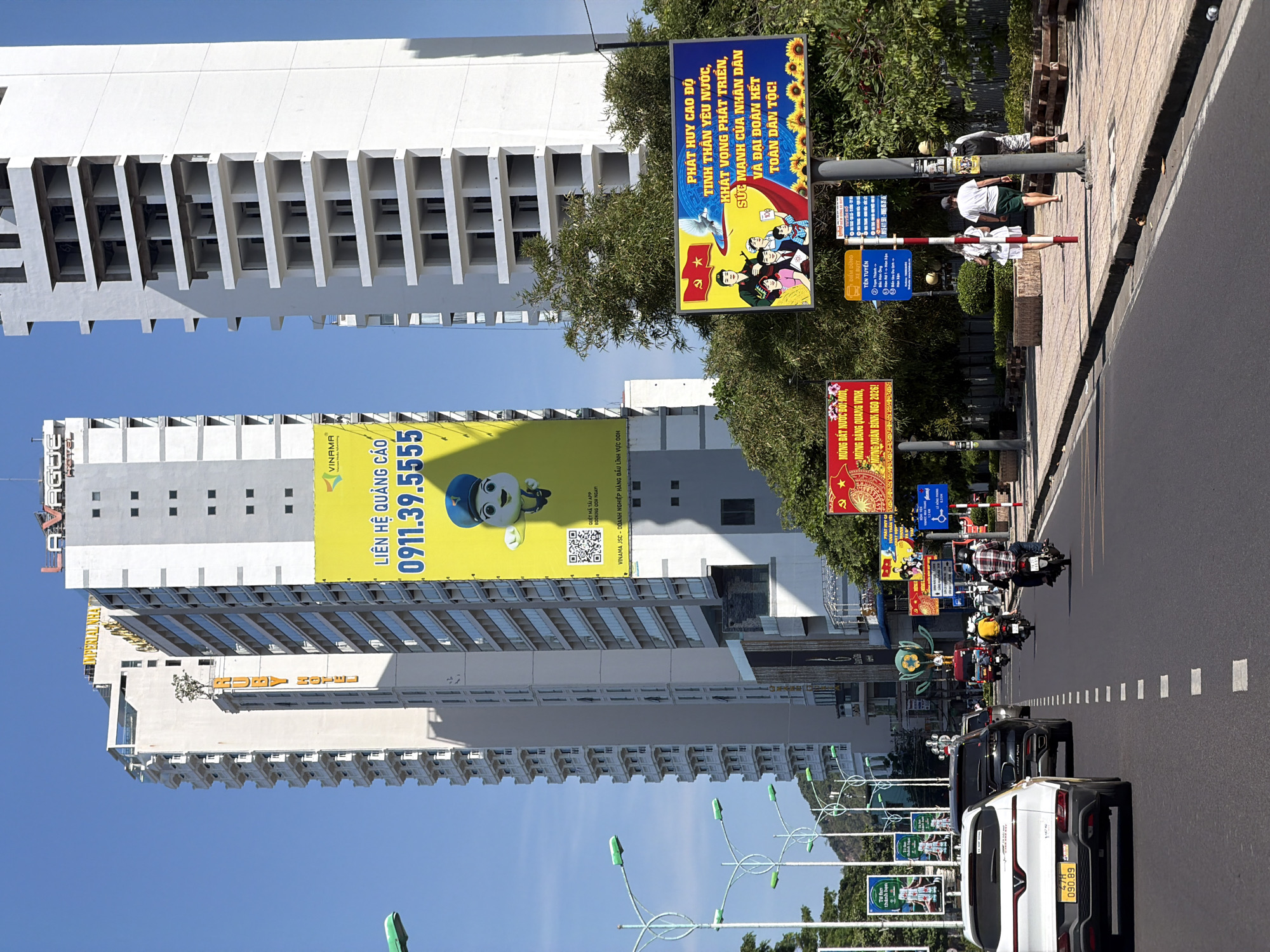The image depicts a street scene in Nha Trang Ward, Vietnam, under a clear blue sky. A multi-lane road dominates the foreground, with vehicles including a white SUV, a black SUV, and several motorcycles in motion. The vehicles are navigating a sunny day, with the road marked with white dashed lines.
On the right side of the road, a sidewalk is visible with several pedestrians. One person is walking away from the camera, dressed in white. Another person, wearing a white t-shirt and green shorts, is walking alongside. Further down, a third person in a backpack is also walking on the sidewalk.
Along the street, there are several large billboards. One prominent billboard is yellow and displays a cartoon character and contact information, "LIÊN HỆ QUẢNG CÁO 0911.39.5555". Another billboard features a political or patriotic message in Vietnamese, depicting a family under a dove and sunflowers. Other smaller billboards are also visible, some with Vietnamese text.
Towering buildings line the street. Two tall, modern buildings are particularly noticeable. One is a white high-rise with visible balconies and windows. The other is a taller, partially constructed building, also white, with exposed floors. The "IMPERIAL Nha Trang" sign is visible on one of the buildings. Streetlights with green lights are spaced along the road. Trees and greenery are present on the sides of the street and between buildings. The overall scene suggests a bustling urban environment in Vietnam.