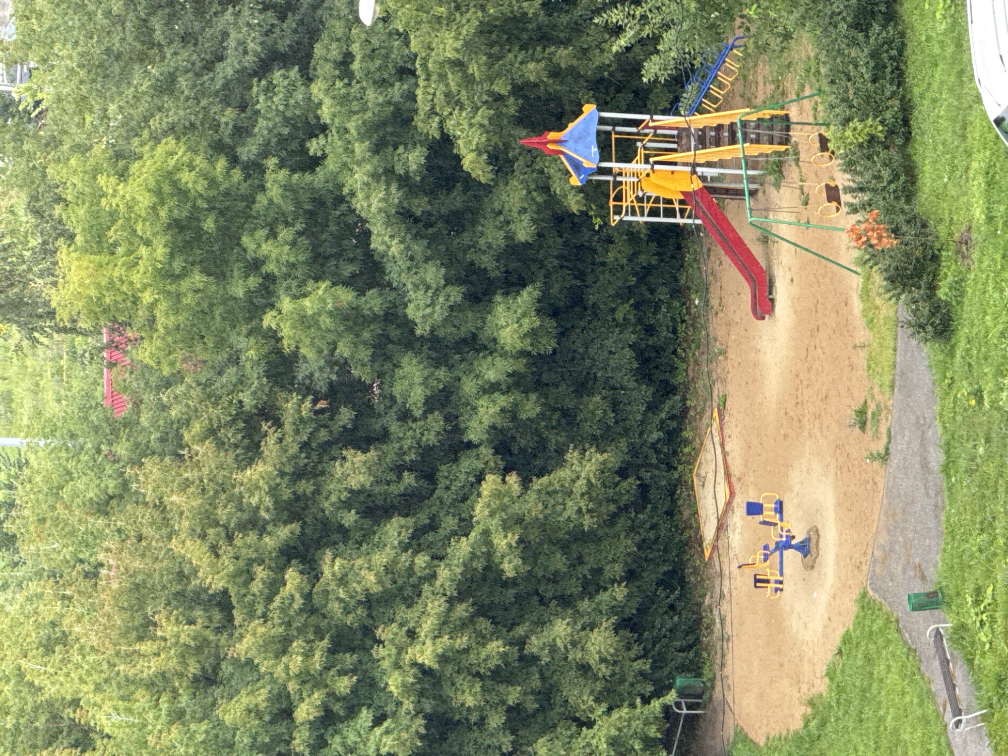 This image depicts an outdoor playground in Nizhny Novgorod, Russia. The playground is situated in a sandy area surrounded by dense green trees. In the center of the sandpit is a blue seesaw with yellow seats. Adjacent to the seesaw is a yellow sandbox with a red frame. To the right of the sandpit, a larger play structure features a red slide, a yellow ladder, and a blue and yellow tower with a red pointed roof. A set of swings with two yellow circular seats hangs from a green frame in front of the play structure. The background is dominated by lush green foliage, suggesting a temperate climate. The overall impression is one of a quiet, somewhat overgrown urban park or residential area.
