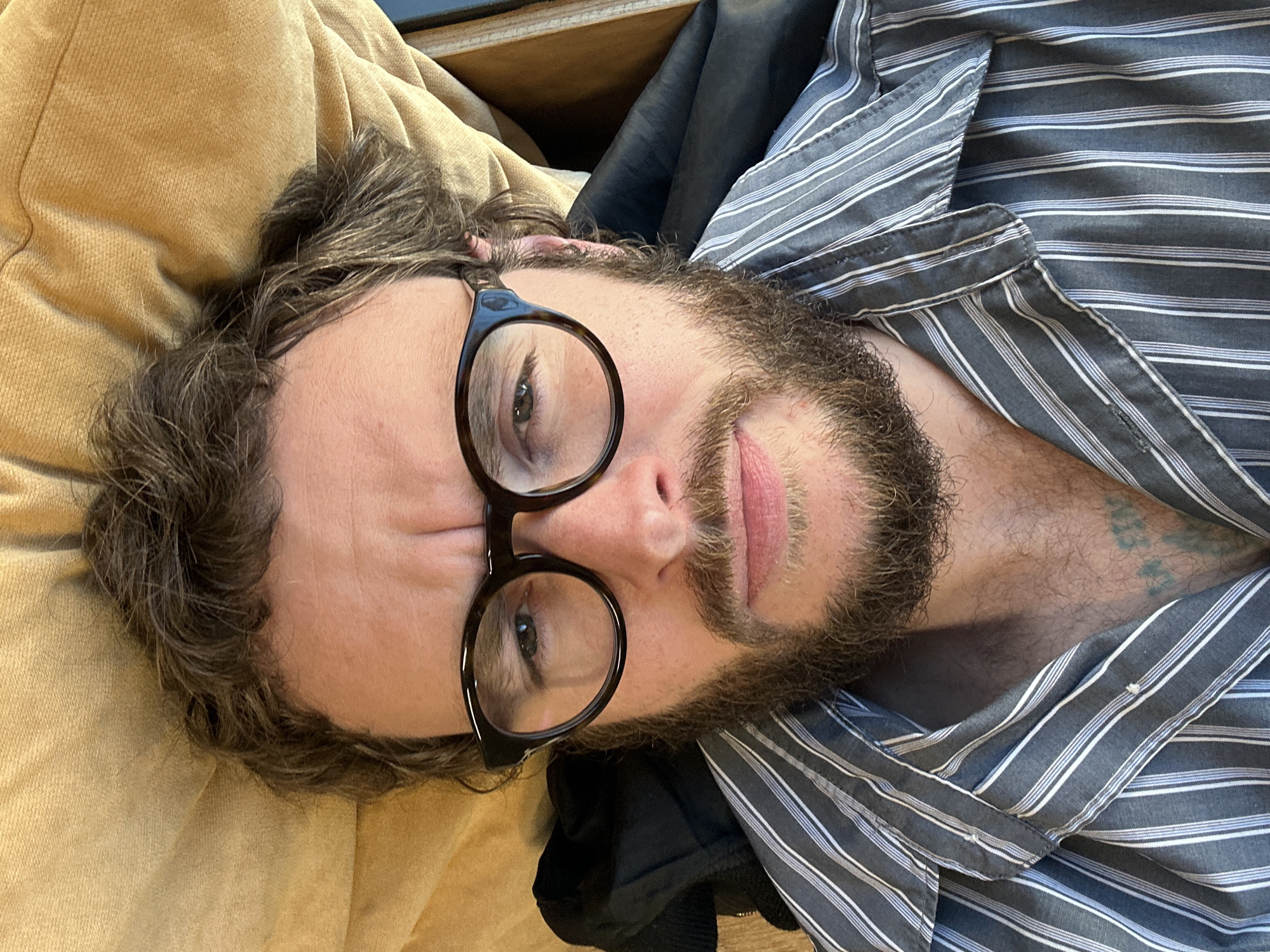 A man is lying down, wearing glasses and a striped shirt. The background includes a tan fabric and a dark, possibly black, garment. No specific location information is discernible from the image.
