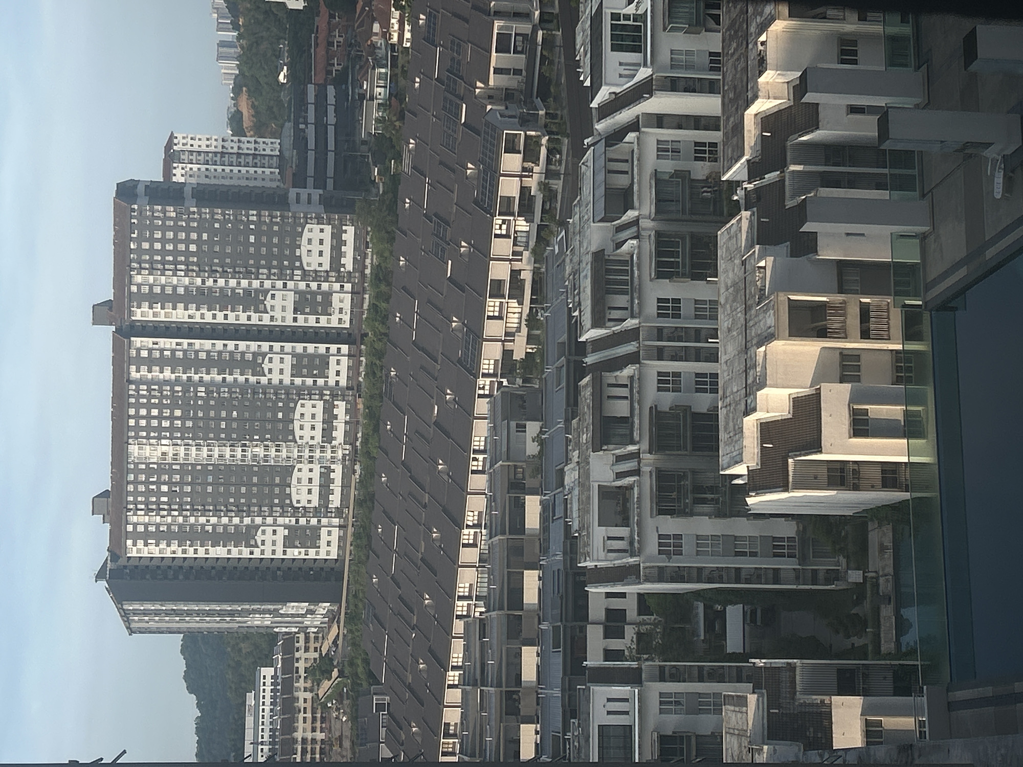 The image presents an aerial view of a densely populated urban area, likely in Kuala Lumpur, Malaysia. The landscape is dominated by a diverse array of buildings, including tall, multi-story apartment complexes, terraced housing with uniform rooflines, and modern, tiered residential buildings with balconies. The buildings are primarily light-colored, featuring white, gray, and beige hues. The architecture is a mix of contemporary and older styles. Interspersed among the buildings are pockets of greenery, suggesting residential landscaping or small parks. The sky is visible in the upper left, appearing clear and light blue. In the lower right corner, there's a blurred, reflective surface, possibly a window or glass barrier, adding a layer of visual complexity to the scene. The overall impression is that of a bustling, densely packed urban environment.