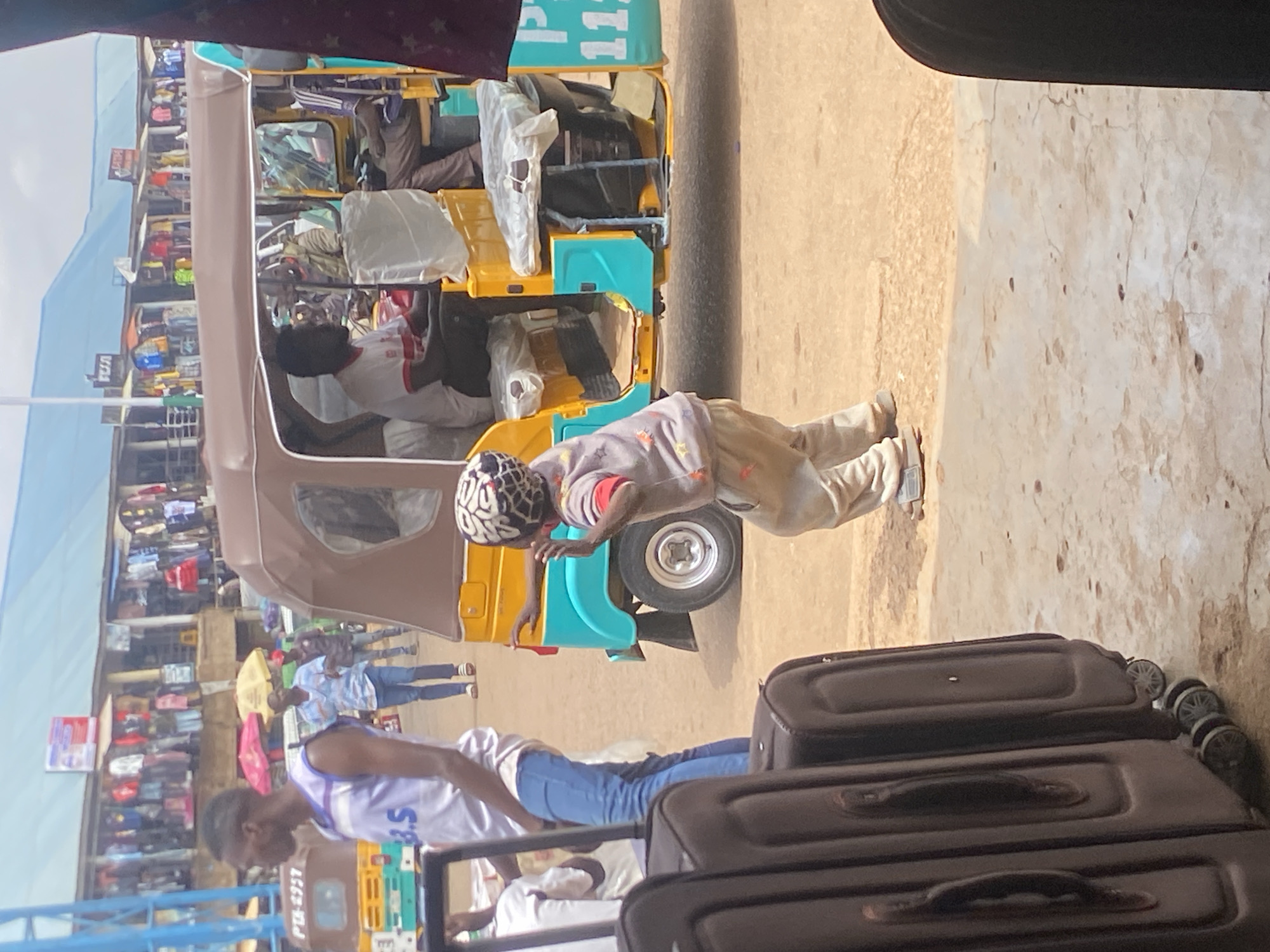 In Bukuru, Nigeria, a young boy wearing a helmet and a light-colored hooded jacket is captured in motion, facing towards a yellow and turquoise auto-rickshaw. He appears to be mid-stride or in the process of a playful movement, with his arms raised. The auto-rickshaw, partially visible, has a brown canopy and contains several passengers. To the left of the scene, two large, dark-colored suitcases are prominently placed. In the background, stalls with clothing displayed are visible under a blue roof structure, suggesting a market environment. The ground is unpaved, with patches of dirt and concrete. A man in a white tank top and blue jeans is standing to the left of the auto-rickshaw, looking towards the right. Several other individuals are present in the background, with one man holding an umbrella and another wearing a red cap. The lighting suggests it is daytime.