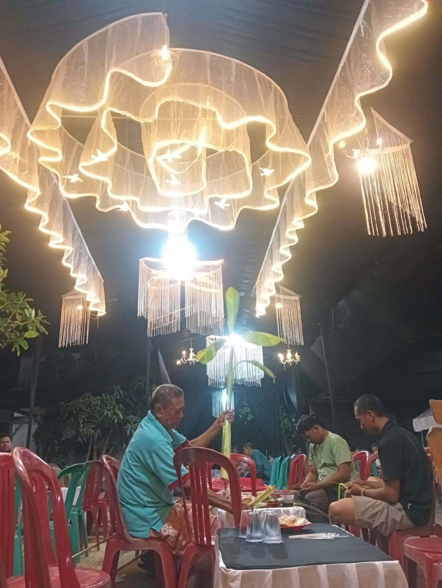 An outdoor evening gathering is taking place under a tent decorated with white, ruffled fabric and string lights that create a wavy effect. Several chandeliers made of white strands hang from the tent ceiling, and a smaller, more traditional chandelier is visible in the background. The scene appears to be in Pucakwangi, Indonesia, likely a community event or celebration given the setup.
In the foreground, three men are seated at a table covered with a black tablecloth. The man on the left, an elderly gentleman wearing a teal shirt and patterned shorts, is holding a leafy green branch. To his right, a younger man in a light green t-shirt and dark pants is looking down, possibly engaged in an activity with his hands. The man on the far right, wearing a black polo shirt and khaki shorts, is also looking down at his hands, which are occupied with what appears to be craftwork involving palm leaves. On the table, there are plates with food, glasses, and cutlery.
Surrounding the table are numerous red and green plastic chairs. In the background, more people can be seen sitting and interacting, suggesting a lively atmosphere. Trees and foliage are visible beyond the tent, indicating the outdoor setting. The overall ambiance is warm and inviting, with the decorative lighting contributing to a festive mood.
