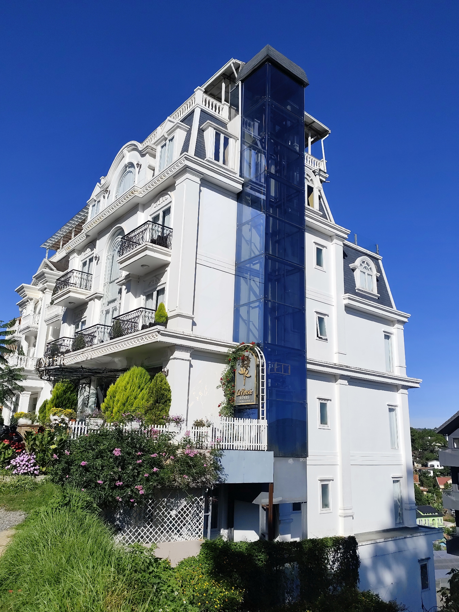 A low-angle, medium-shot captures a five-story, white hotel under a clear blue sky. The hotel features a mansard roof, intricate wrought-iron balconies, and a modern glass elevator shaft extending up one side. At the base of the hotel, a white picket fence encloses a garden with various plants and flowers in bloom. Lush green grass covers the foreground, adding to the natural setting. In the background, other buildings are visible, nestled amongst trees on a hillside. A sign near the entrance identifies the building as "La Pensée Hotel." This hotel is located in the city of Da Lat, Vietnam. There are no people visible in the image.
