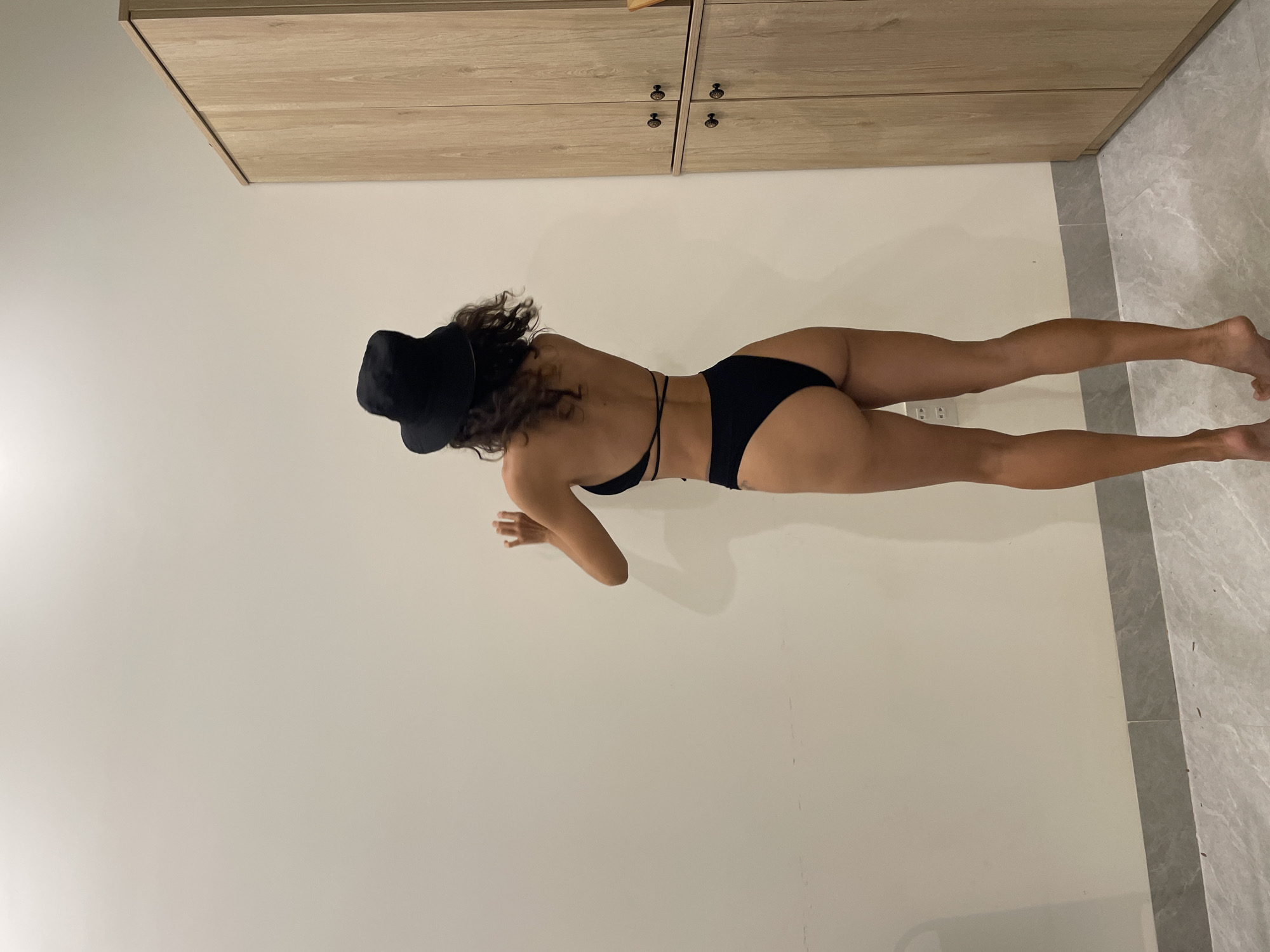 An indoor photograph, taken in Mũi Né, Vietnam, shows a woman lying prone with her back to the camera. She wears a black two-piece swimsuit, featuring a strappy top, and a black hat that covers most of her curly brown hair. Her body is athletic, and she rests on her forearms, with her legs extended straight behind her.
The setting is a minimalist room. The surface she lies on is a light, plain color, possibly an off-white floor or mat. To the right, the floor transitions into grey marbled tiles. An electrical outlet is visible on the light-colored wall near her left leg. Above her, a light-colored wooden cabinet or wardrobe with two pairs of dark handles is mounted on the wall. The lighting suggests a bright indoor environment. The woman's pose conveys a relaxed state. No visible text is present.