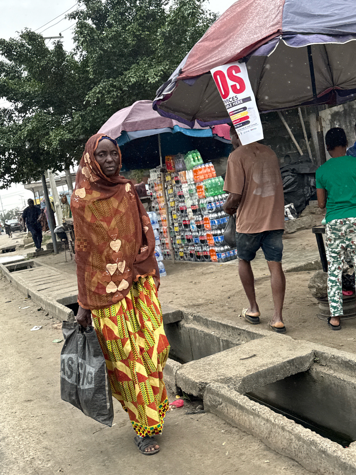 This image captures a street scene in Ado, Nigeria. In the foreground, a woman is walking, carrying a dark-colored shopping bag in her left hand. She is wearing a patterned headscarf and a colorful skirt with geometric designs. To her right, a man in a brown t-shirt and dark shorts is walking away from the camera, holding a dark bag. He has a sign with red lettering that reads "OS SERVICES AVAILABLE HERE" attached to his head, under an umbrella. Behind him, another person in a green shirt and patterned pants is visible. The background features stalls with stacks of bottled beverages and other goods, suggesting a marketplace or roadside vendors. The environment appears to be an outdoor setting with a paved walkway and a concrete drainage ditch. The sky is overcast, and there appear to be light rain or water droplets in the air.
